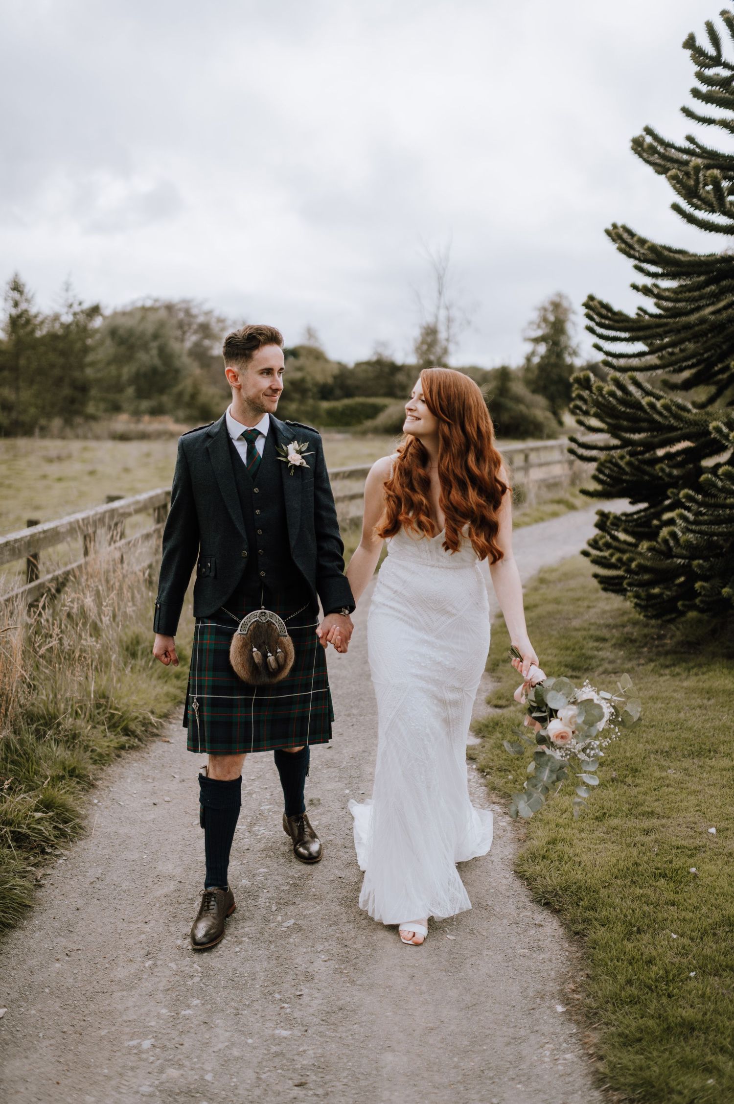A couple walks along a countryside path during their Scottish wedding, with the groom in traditional kilt.