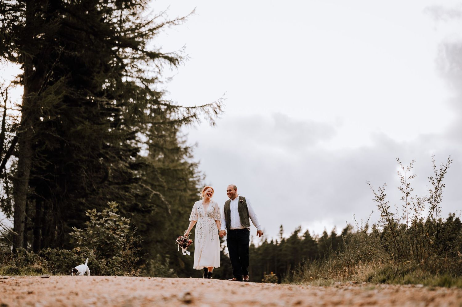 A couple walks along a forest path with their small dog on a cloudy day.