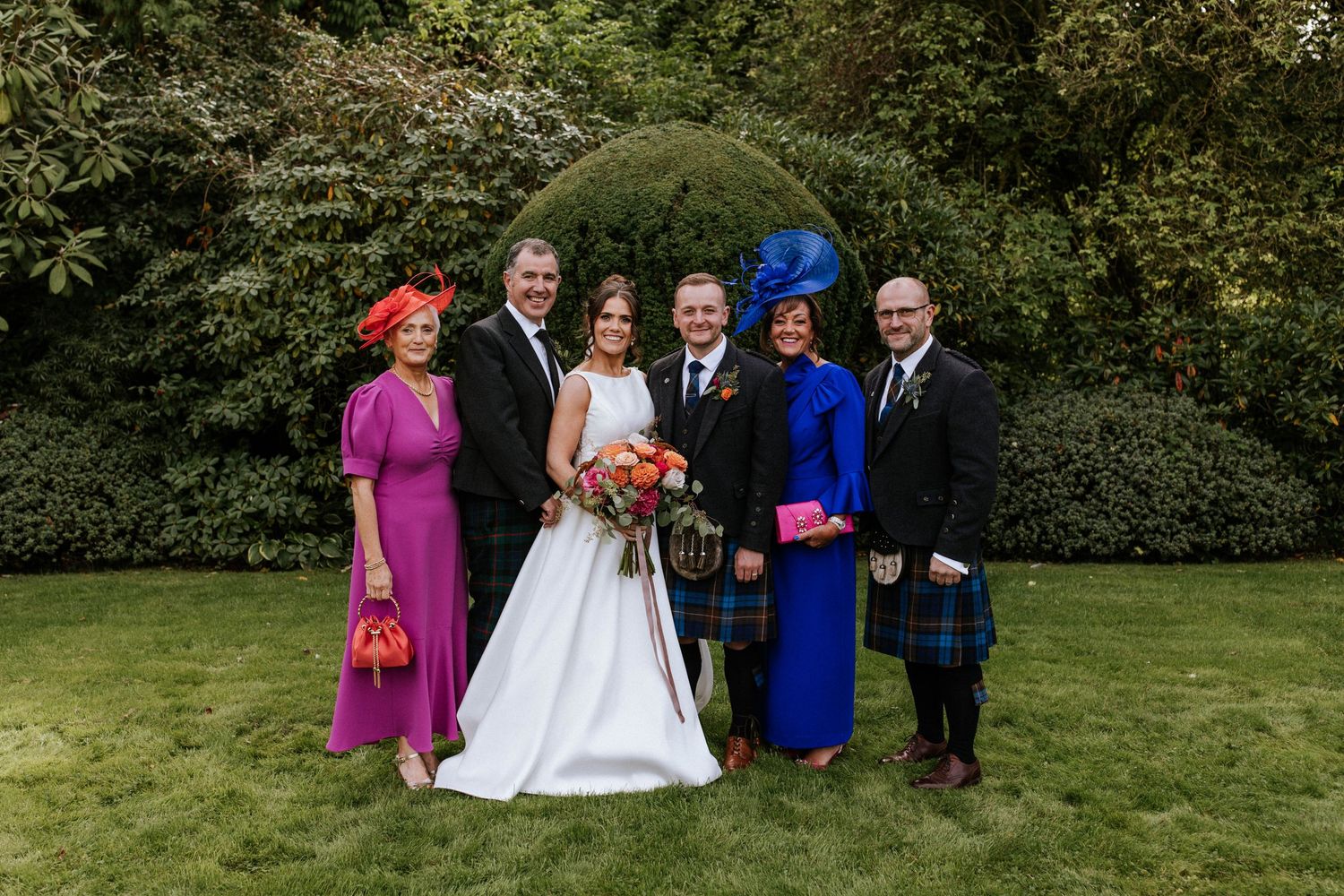 Formal wedding group photo taken outdoors with greenery backdrop and traditional Scottish elements.