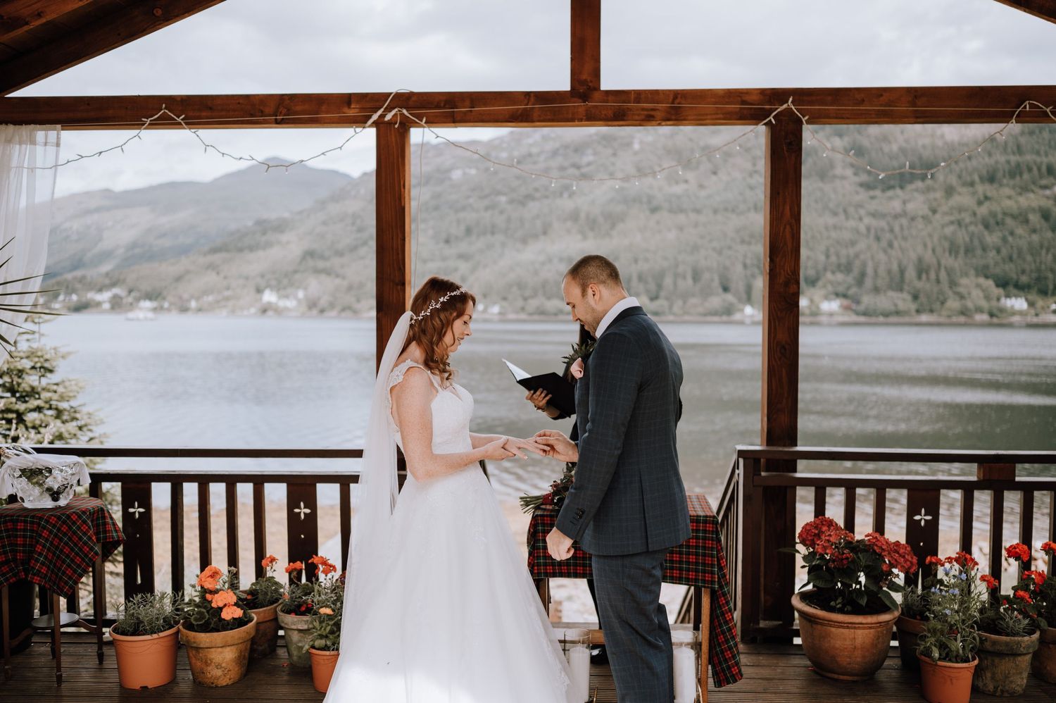 Couple exchanges vows under a wooden gazebo overlooking a serene mountain lake with potted flowers on the railing.