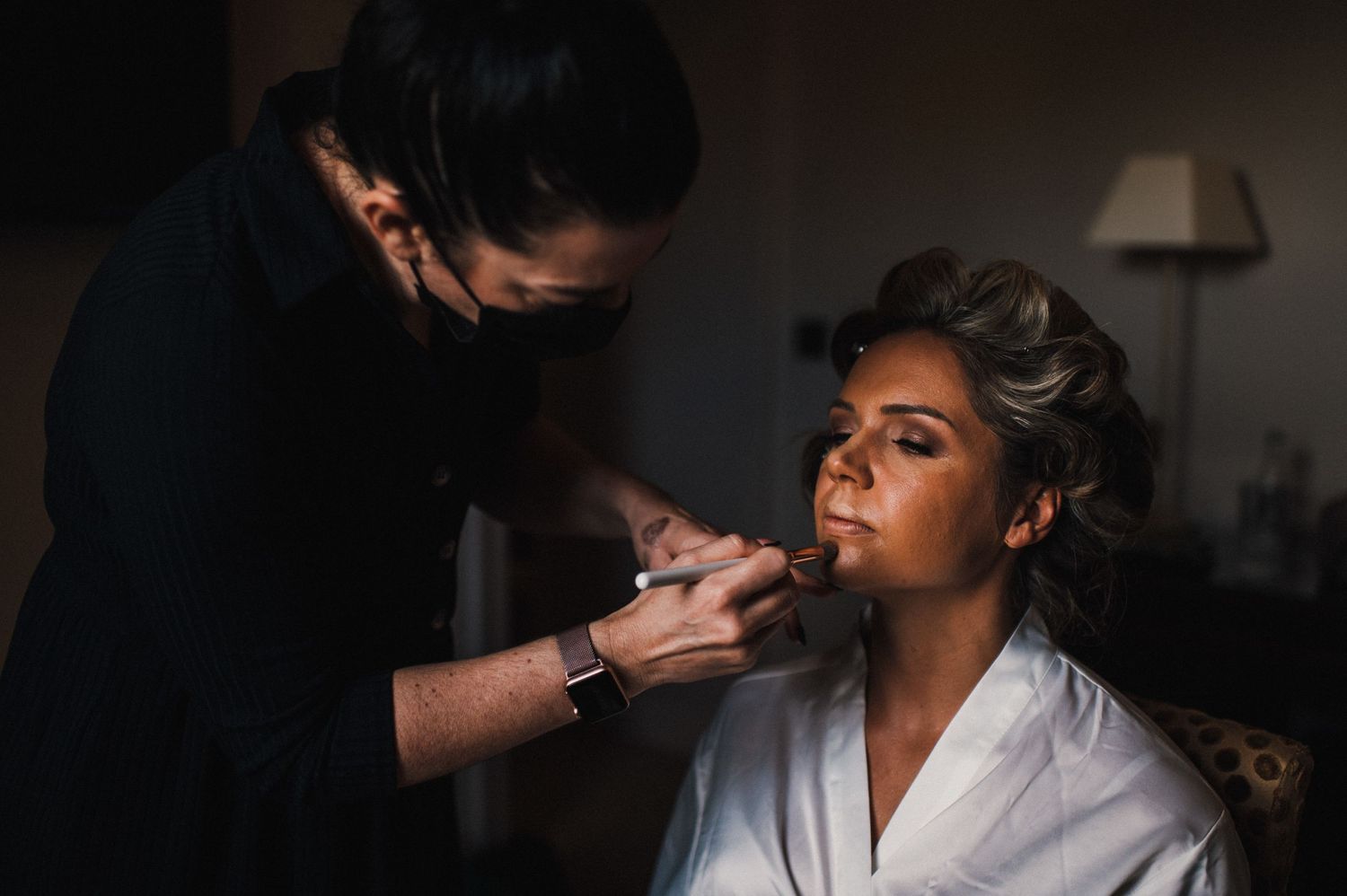 Makeup artist applying cosmetics in a dimly lit room during wedding preparation.
