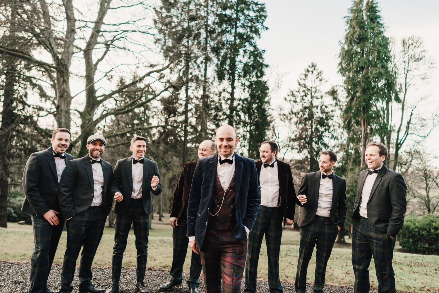 Groomsmen in black tie attire stand together for a group photo in a wooded setting.