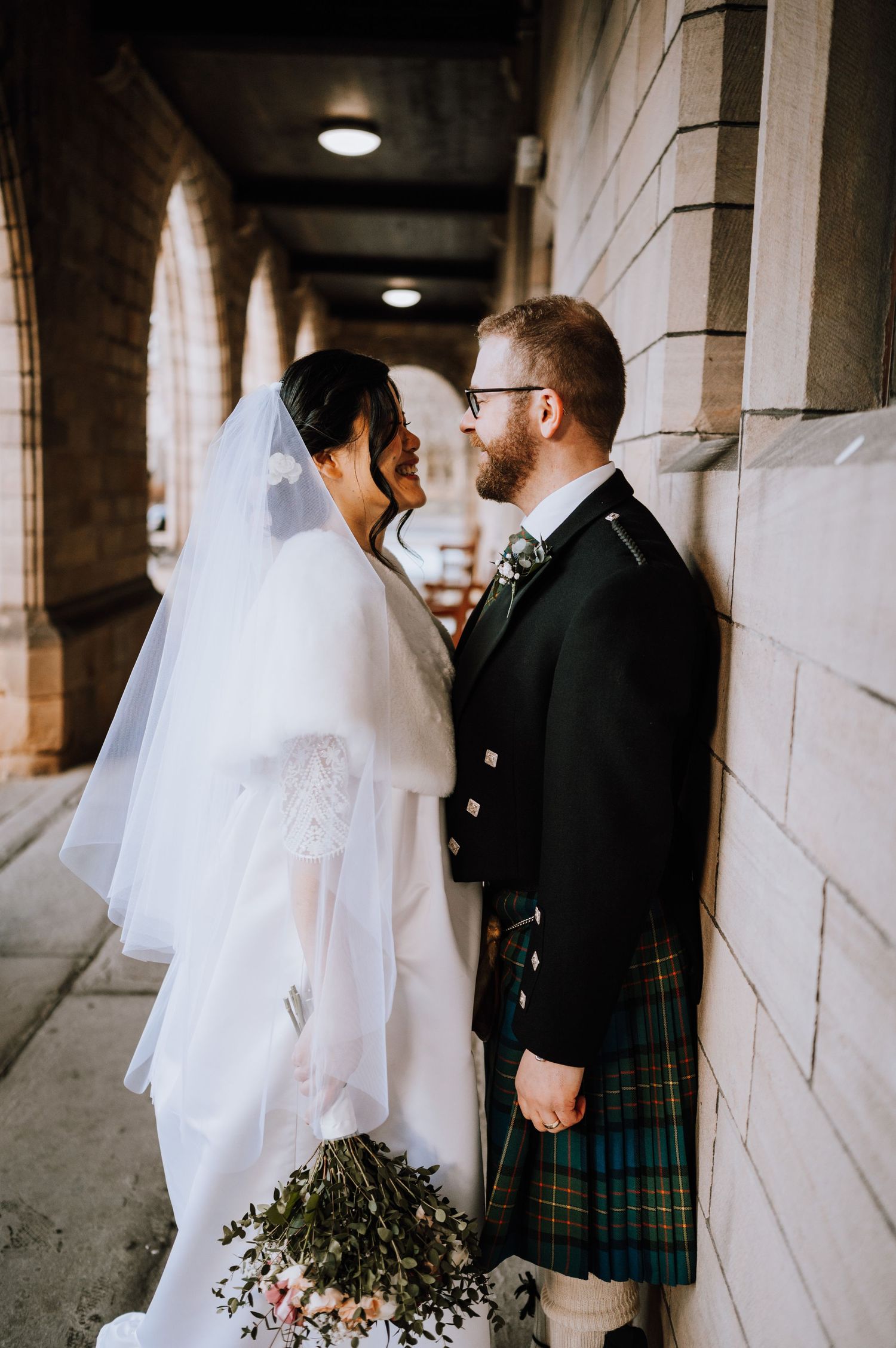 Newlyweds share intimate moment in church corridor with groom wearing traditional Scottish kilt.