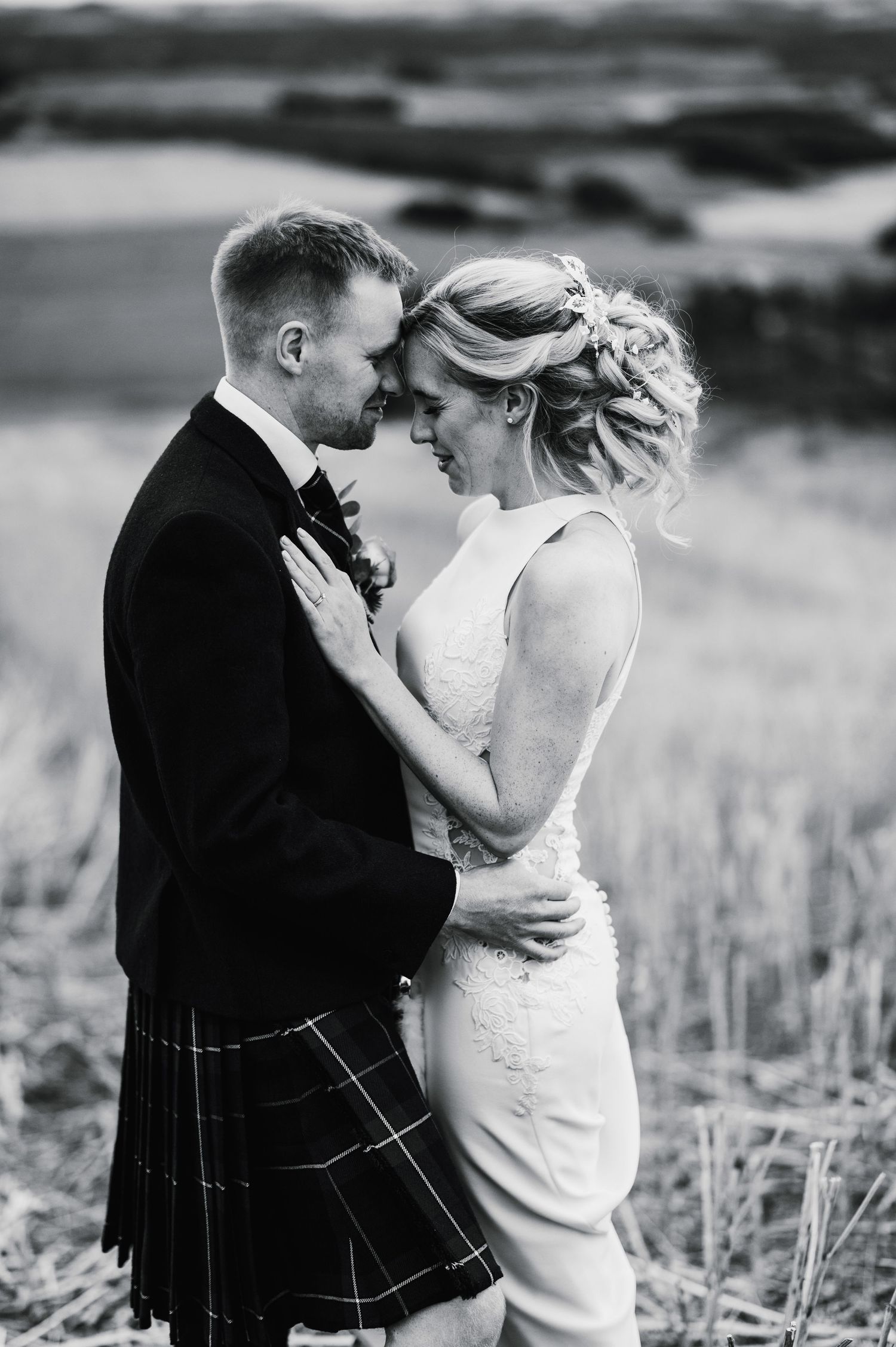 Black and white portrait of a romantic wedding couple embracing in Scottish highland attire against a rural landscape.
