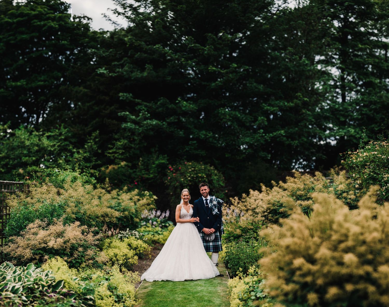 Wedding couple walking through a lush garden pathway surrounded by blooming flowers and greenery.