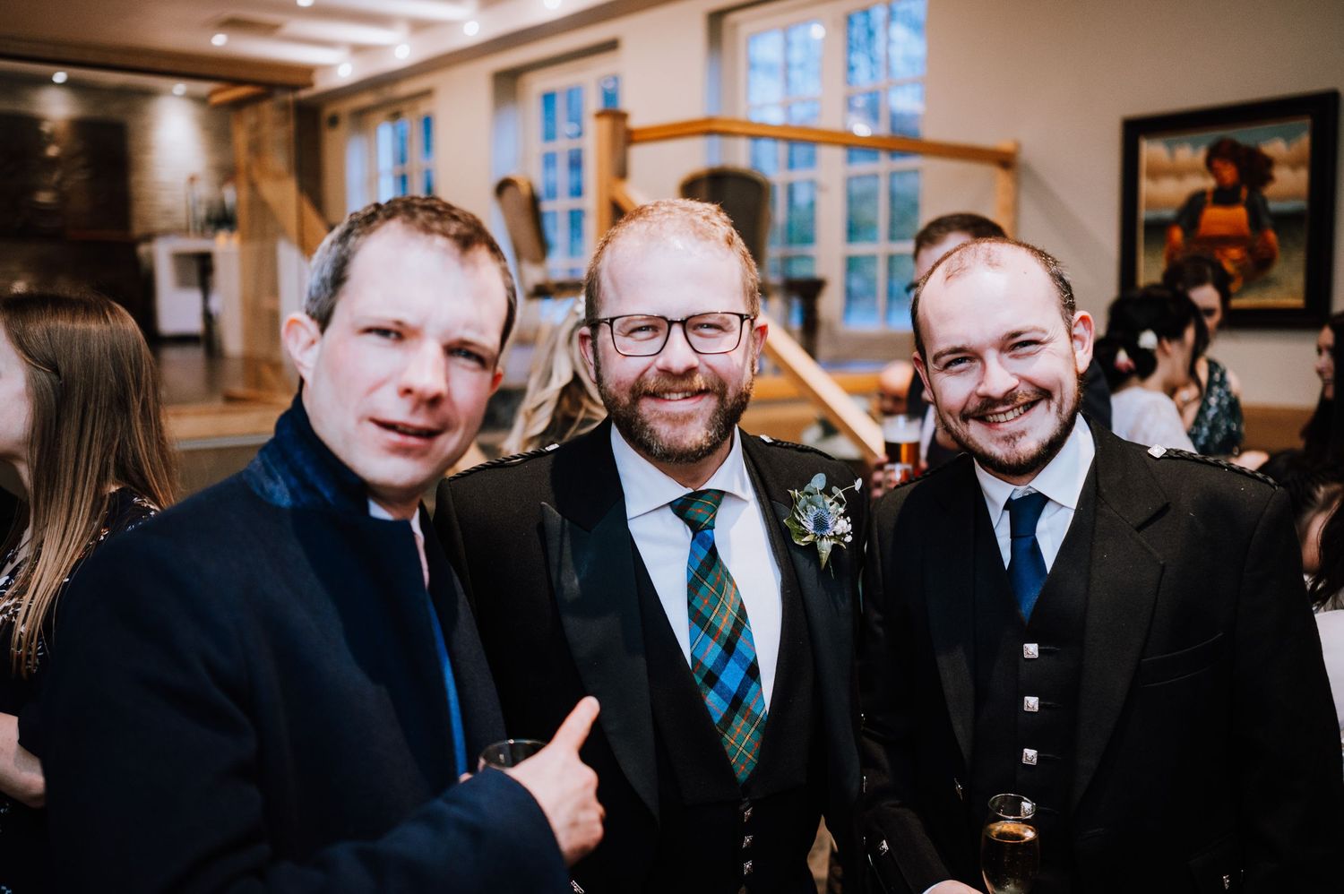 Three guests in formal wear smile together at an indoor wedding reception.