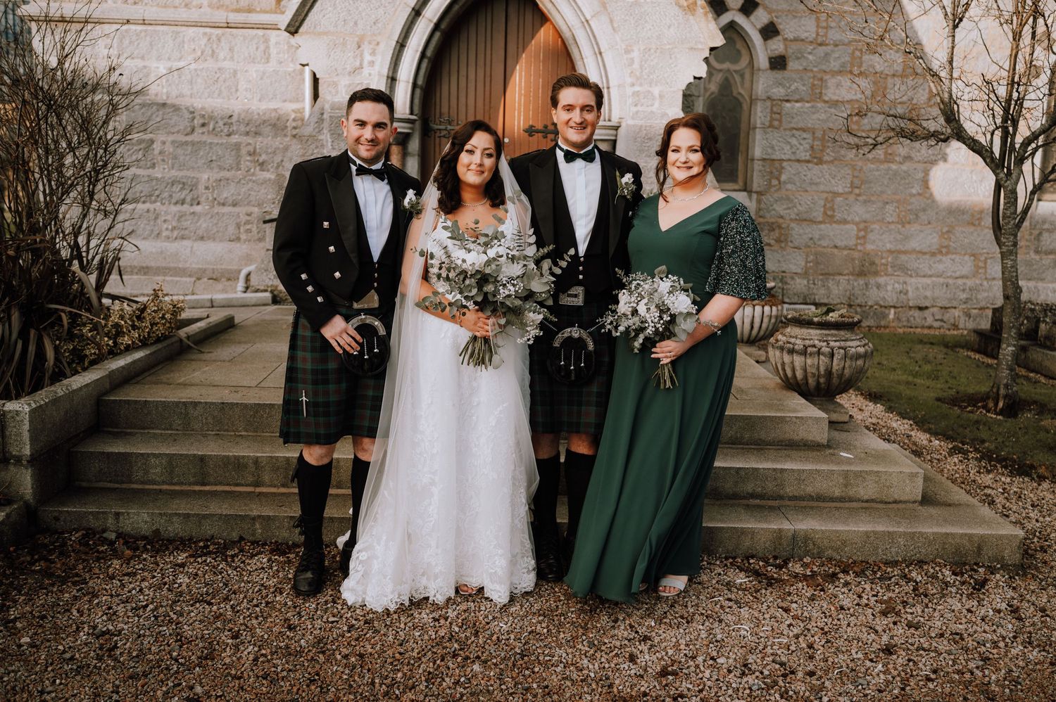 Wedding party in traditional Scottish attire with kilts and emerald green dresses pose on church steps.
