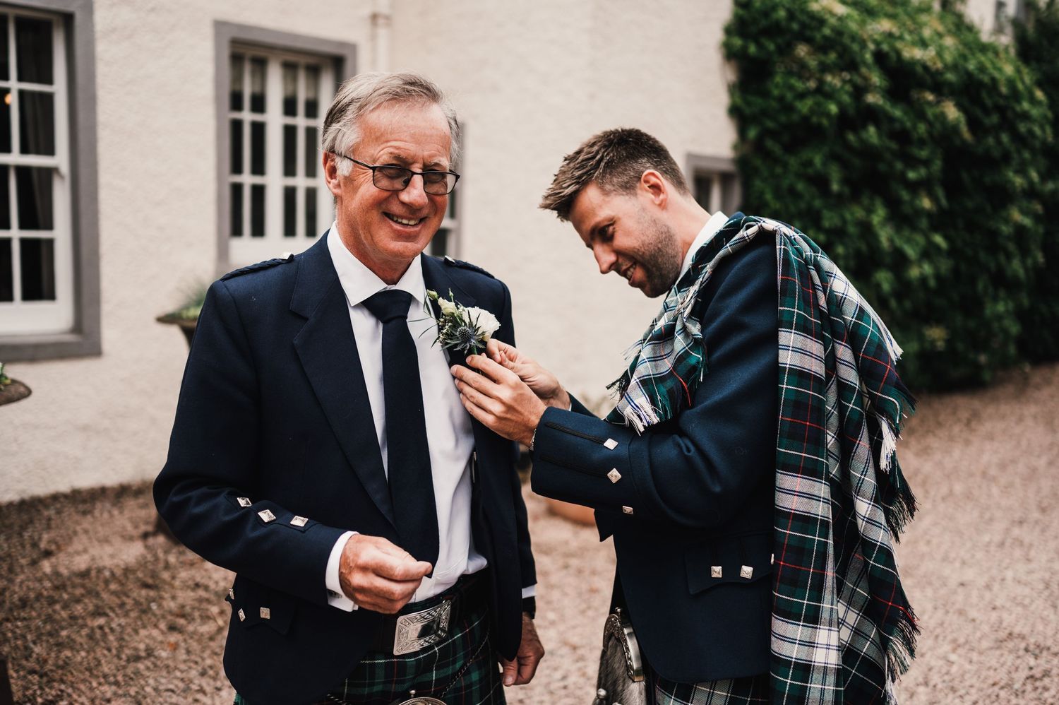 Two gentlemen in traditional Scottish attire share a lighthearted moment while holding beverages.