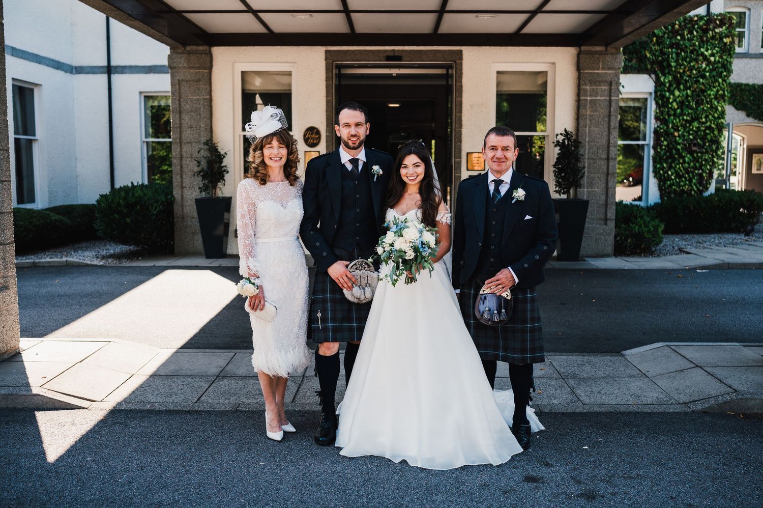 Wedding party poses formally in traditional Scottish attire outside a elegant venue entrance.