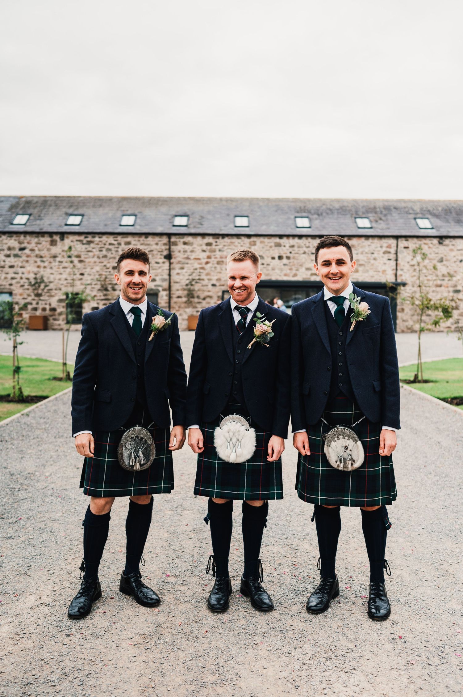 Three groomsmen in matching kilts stand on a gravel path in front of a rustic venue.