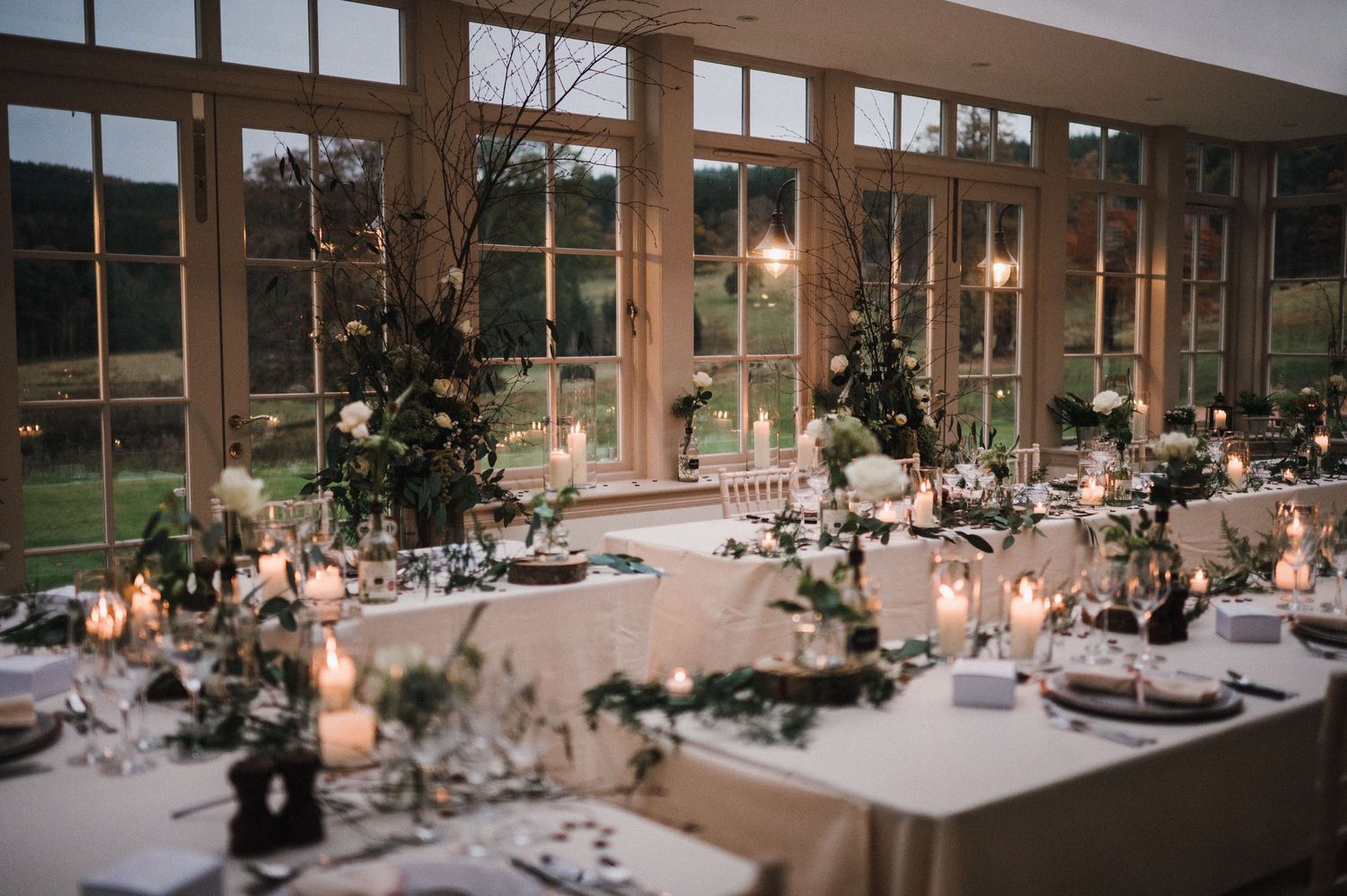 Wedding reception room with large windows and elegant table settings at dusk.