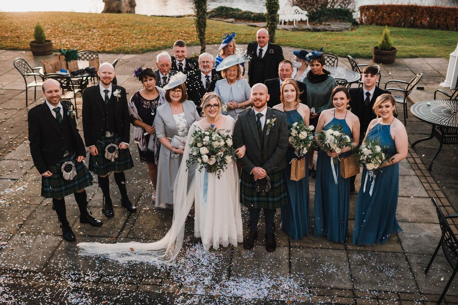 Scottish wedding party poses together outdoors with men in traditional kilts and women in blue dresses.