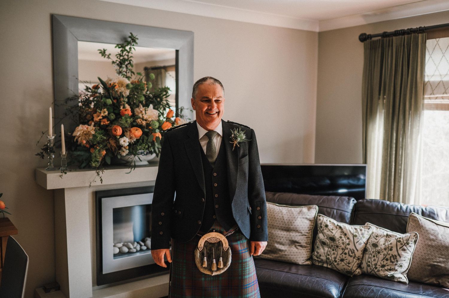 A person wearing a traditional dark kilt suit stands in an elegant living room with floral arrangements.