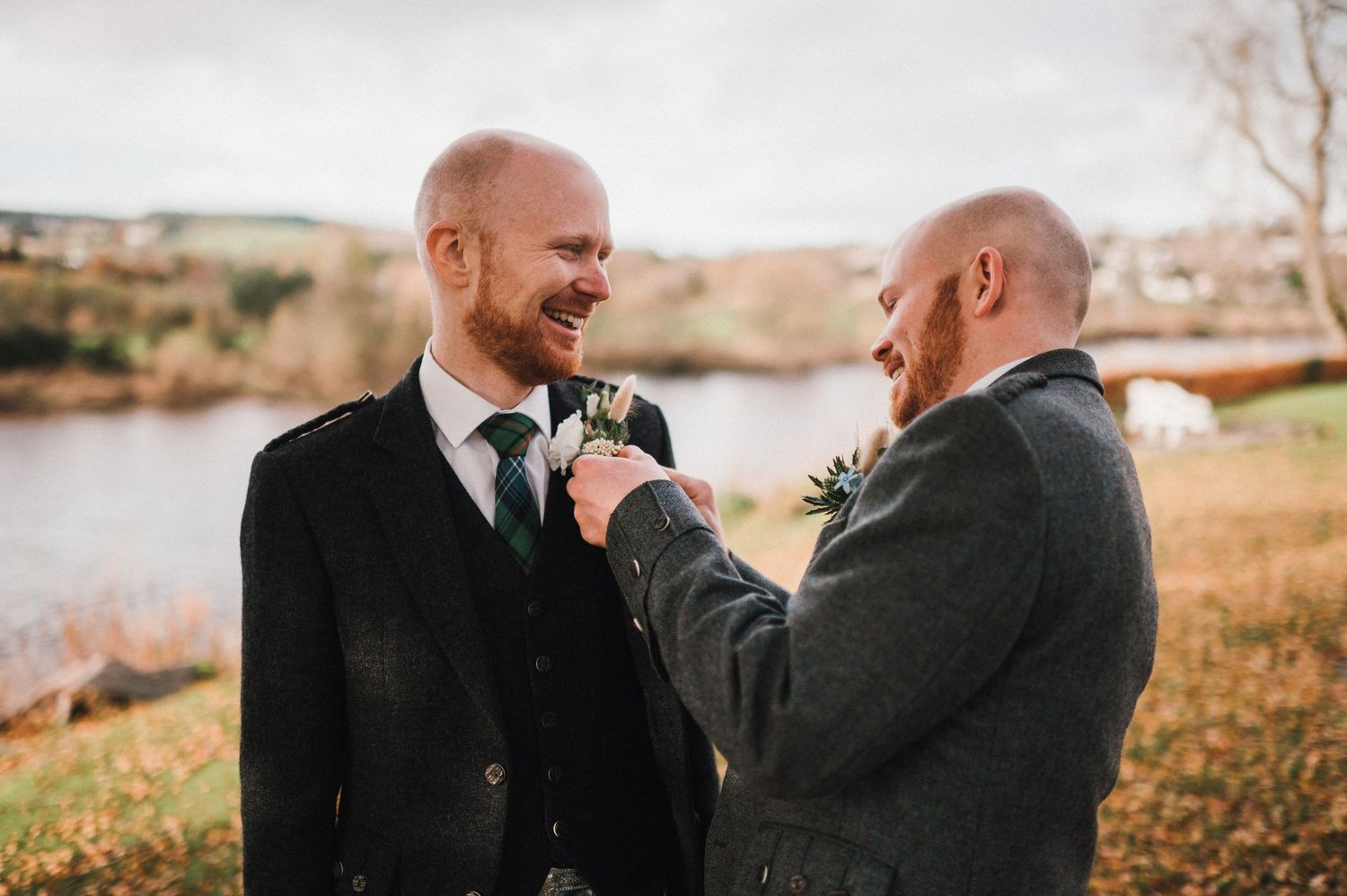 Two people in dark suits laughing while fixing boutonnieres outdoors.