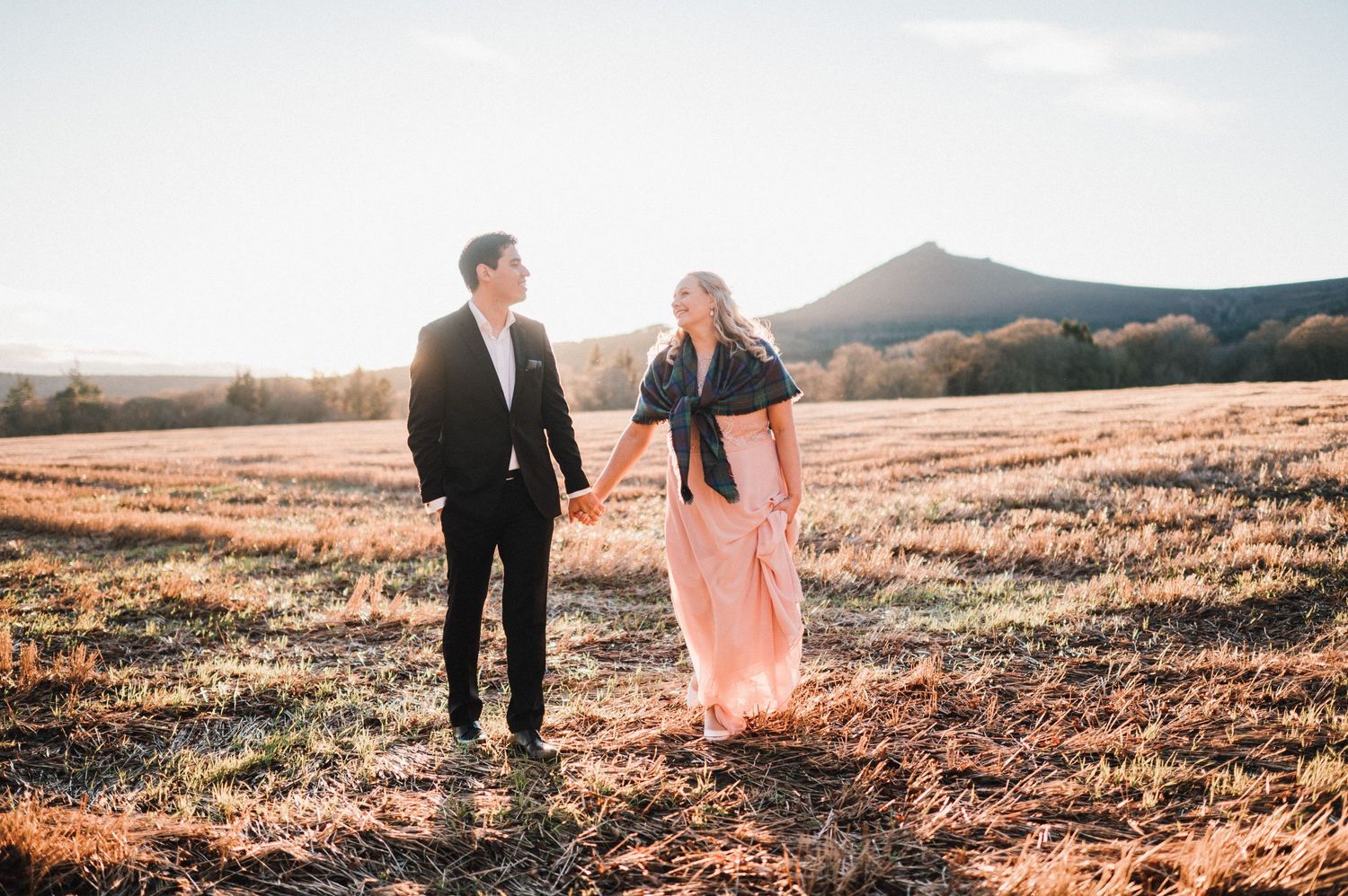 A couple stands together in an open field during golden hour with mountains in view.