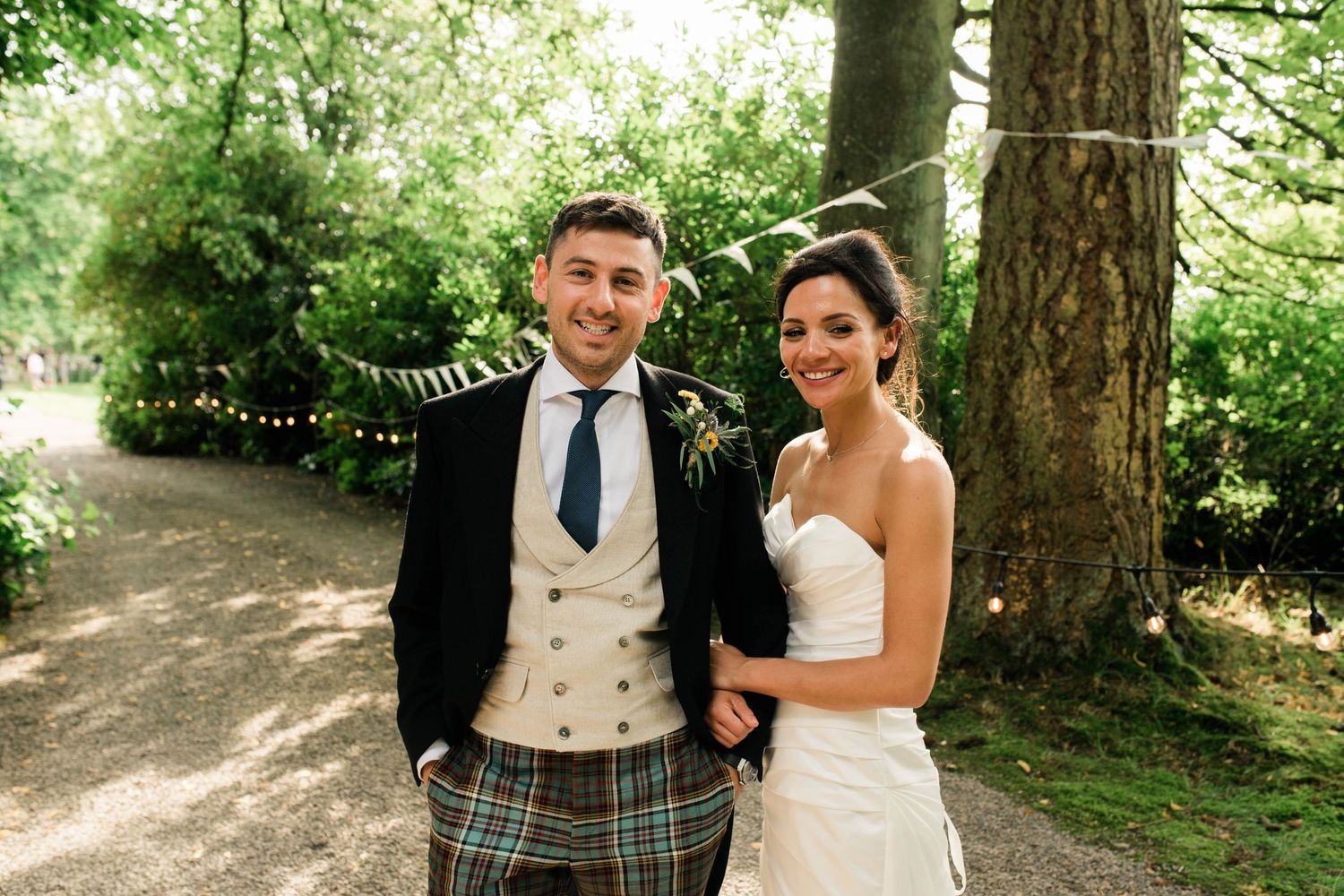 A Scottish wedding couple poses outdoors on a forest path with the groom in traditional highland dress and tartan kilt.