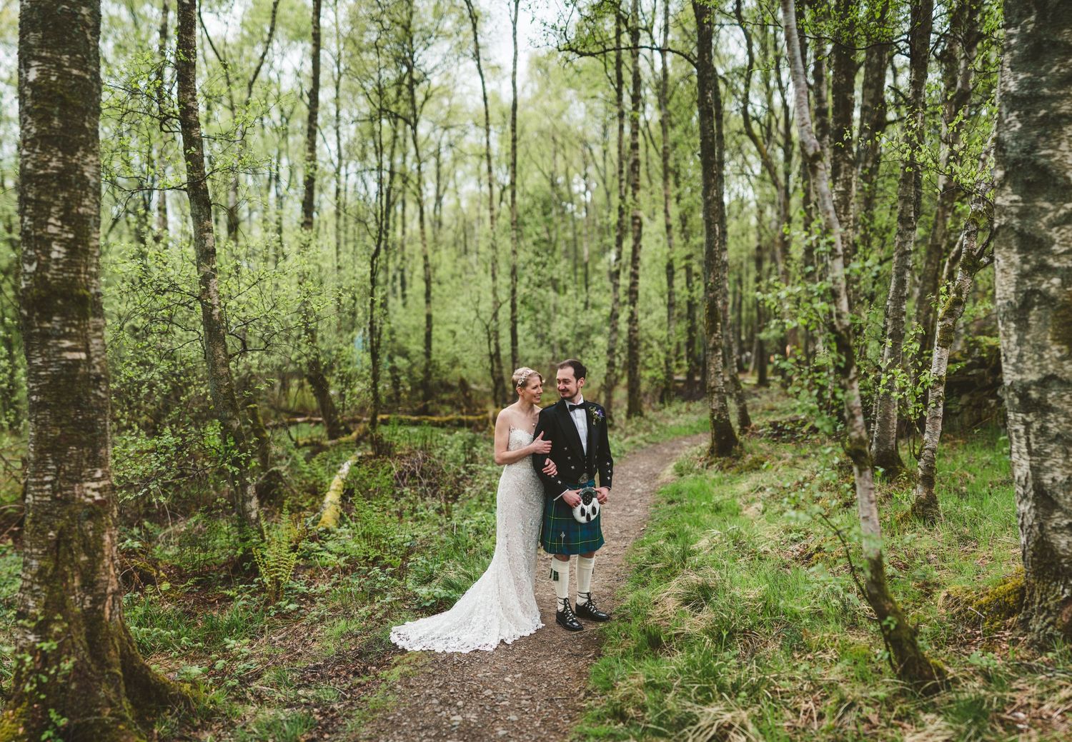 A couple in wedding attire walks through a verdant spring forest on a natural path.