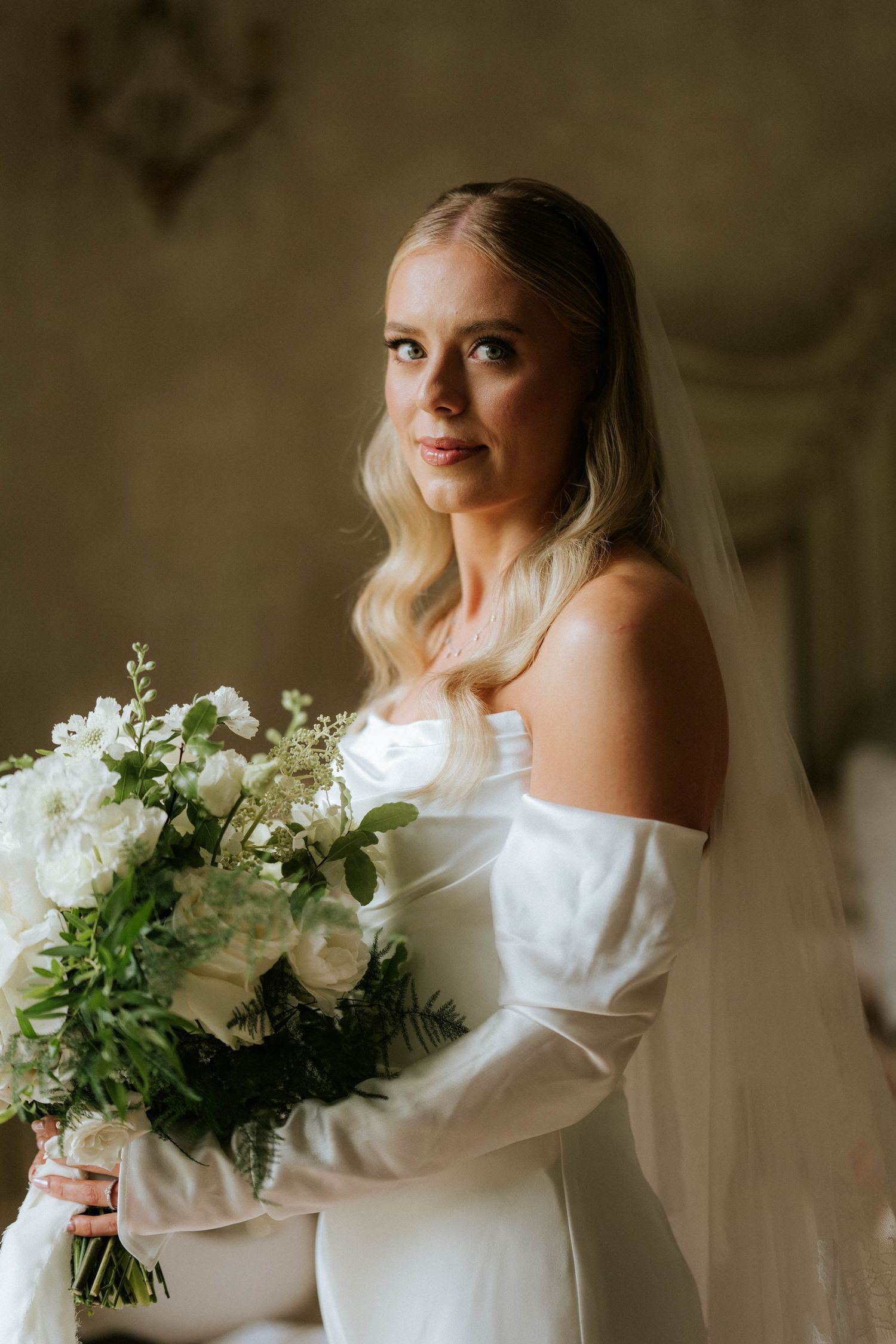 Bride in elegant long-sleeved white gown holds lush white and green bouquet in romantic indoor setting.