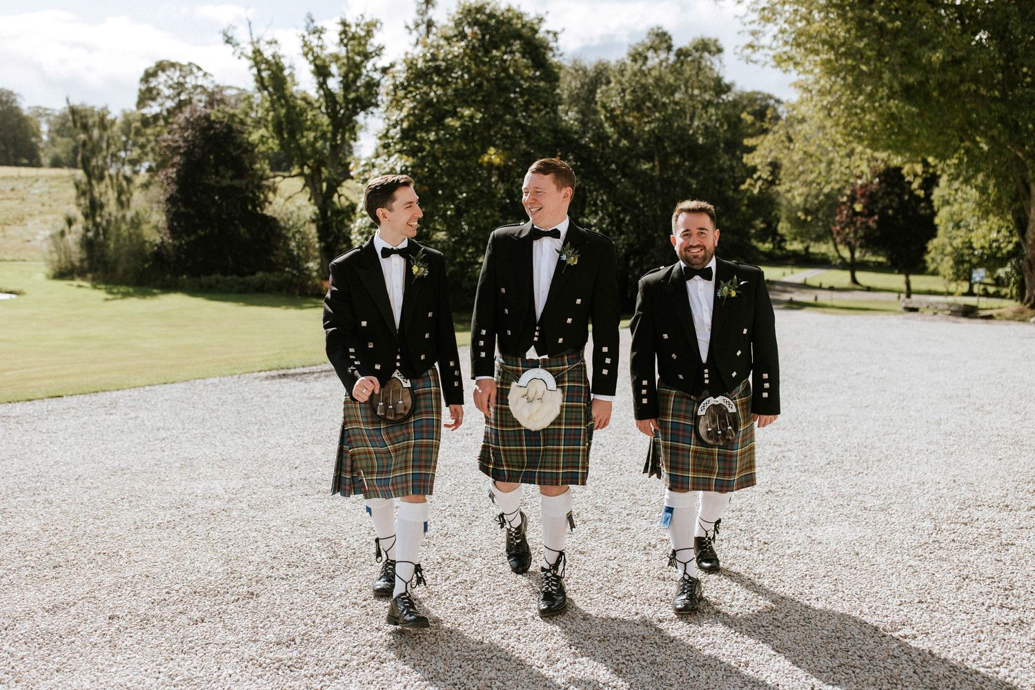 Groomsmen in traditional Scottish kilts walk along a gravel path at an outdoor wedding venue.