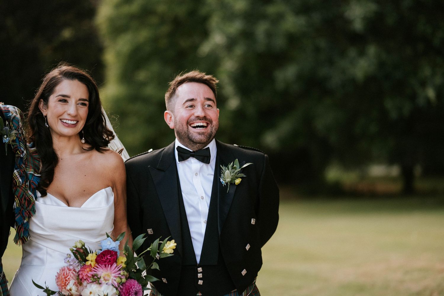 A joyful wedding couple laughs together outdoors while holding a vibrant pink and purple bouquet.