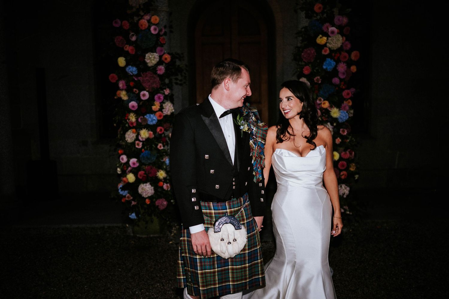 A couple poses against floral decorations, with the groom wearing traditional Scottish formal attire.