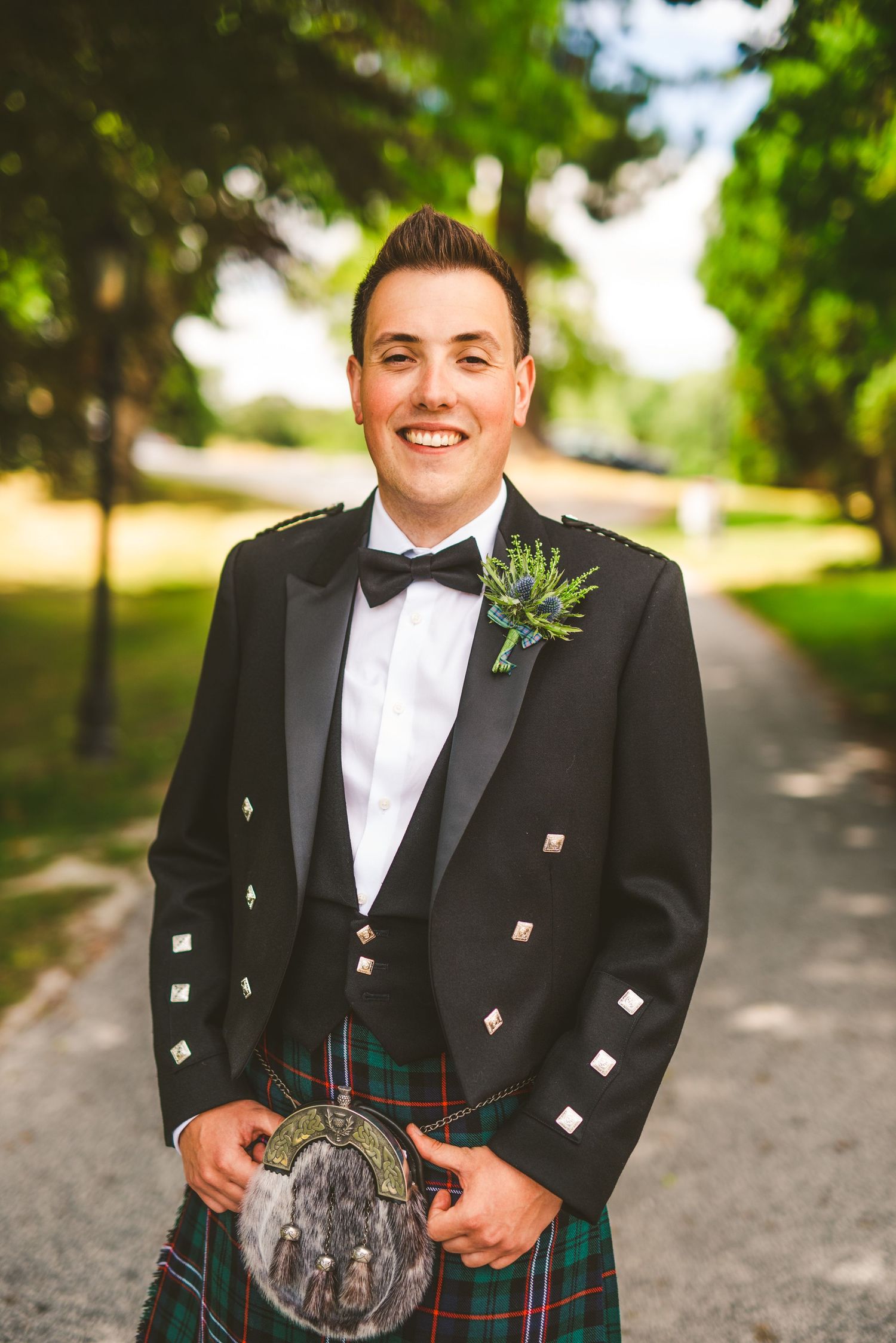 A person wearing traditional Scottish formal attire including a black jacket and kilt stands on a tree-lined path.
