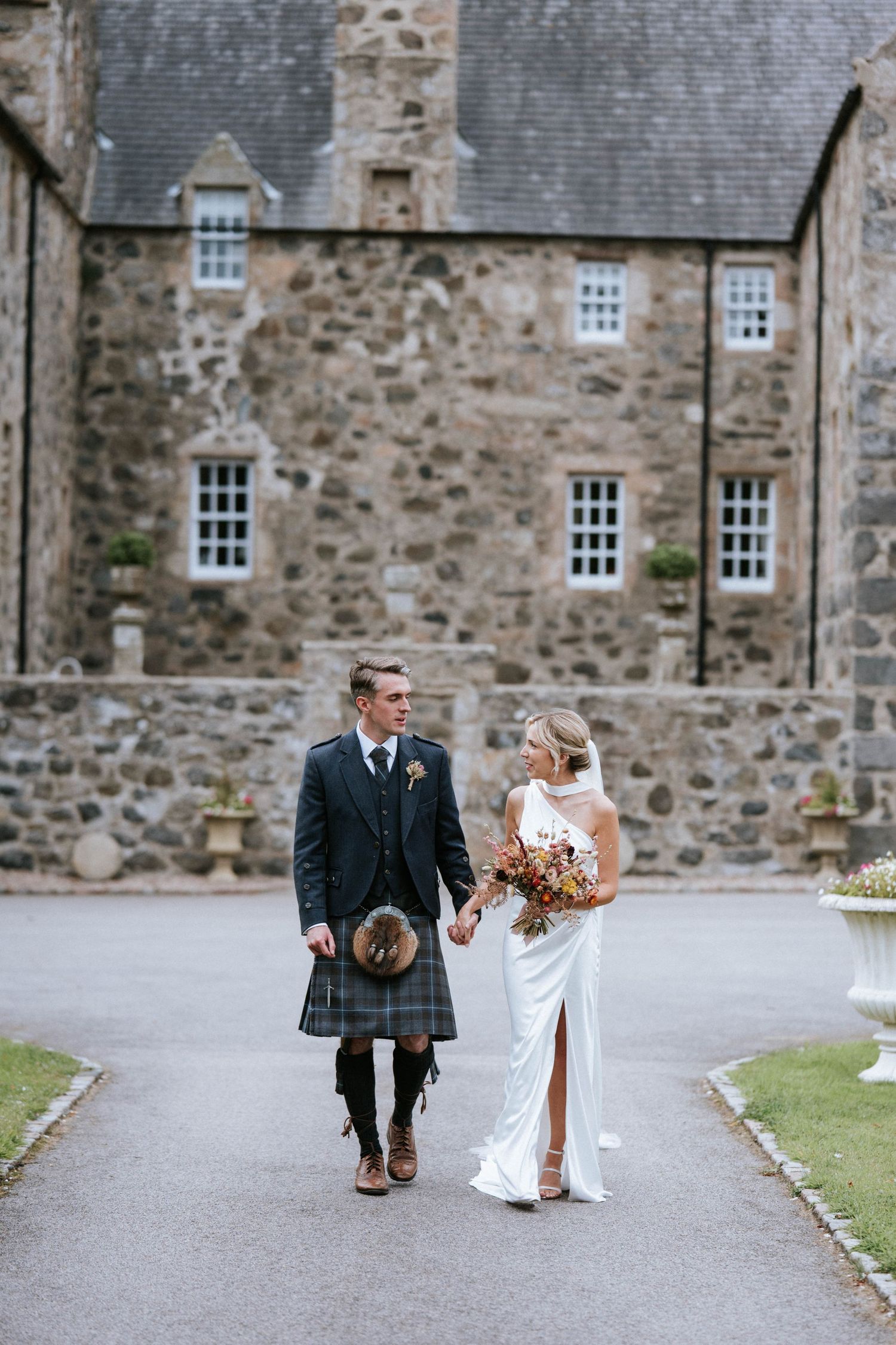 A couple walks together on a path outside a stone castle, with the groom wearing traditional Scottish kilt attire.