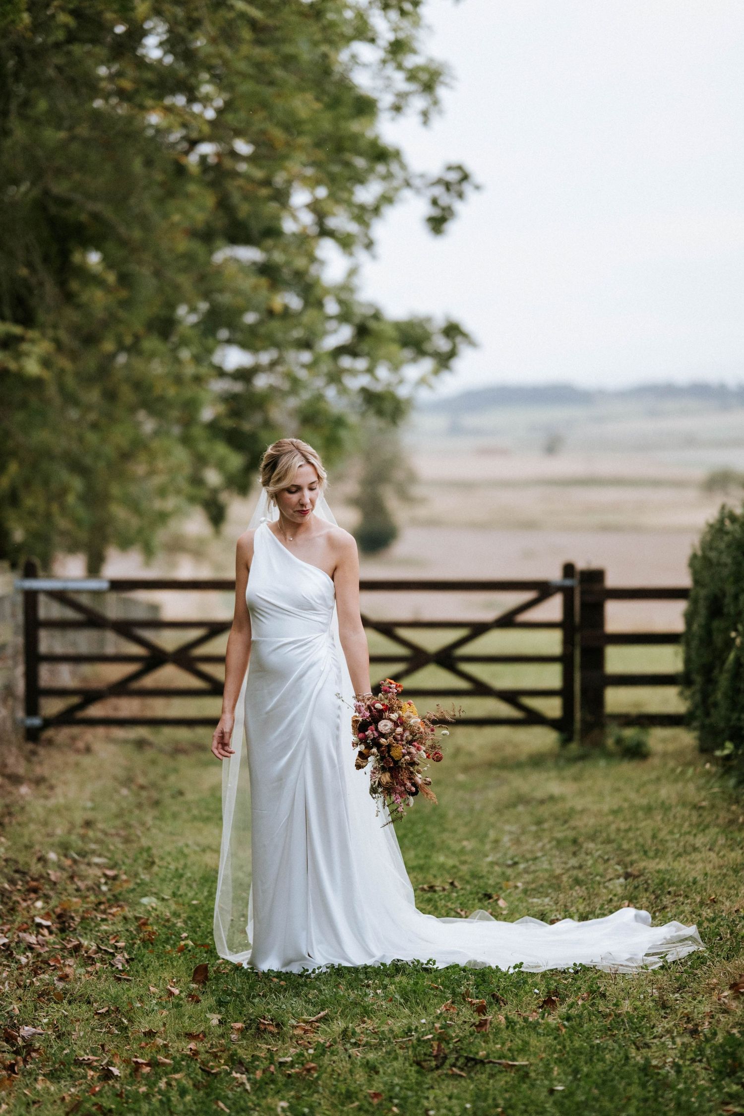 A bride in an elegant one-shoulder white gown poses in a rustic countryside setting with a wooden gate and fields behind.