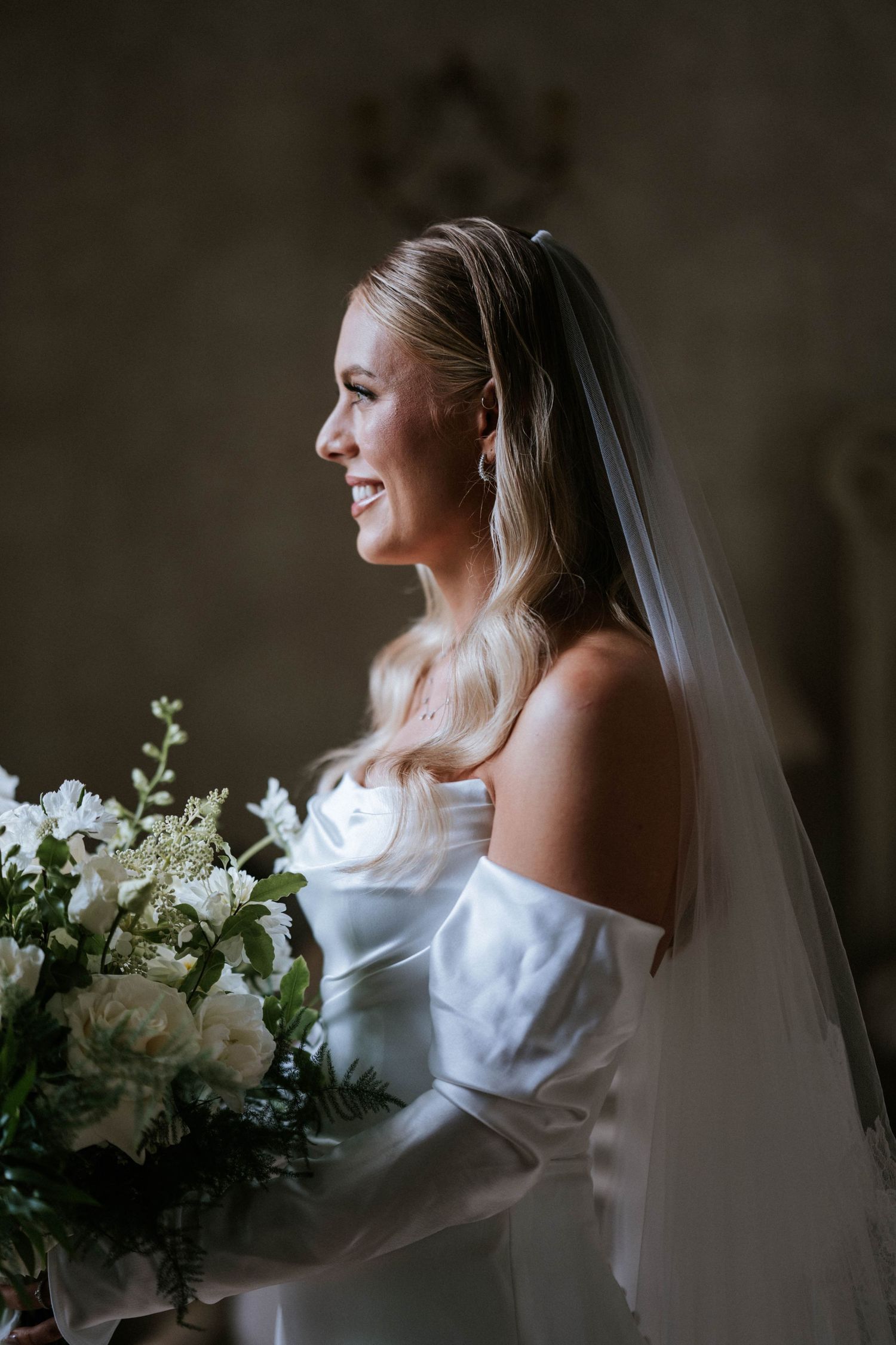 Bride in white dress holding bouquet poses in dramatic window light.