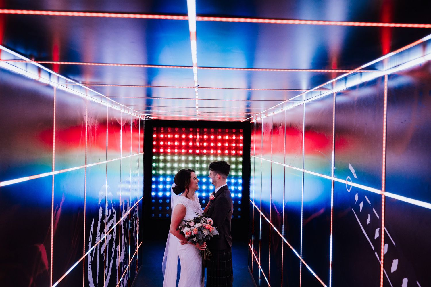 Performers dance in a futuristic corridor with neon lighting and illuminated grid patterns on stage during a show.