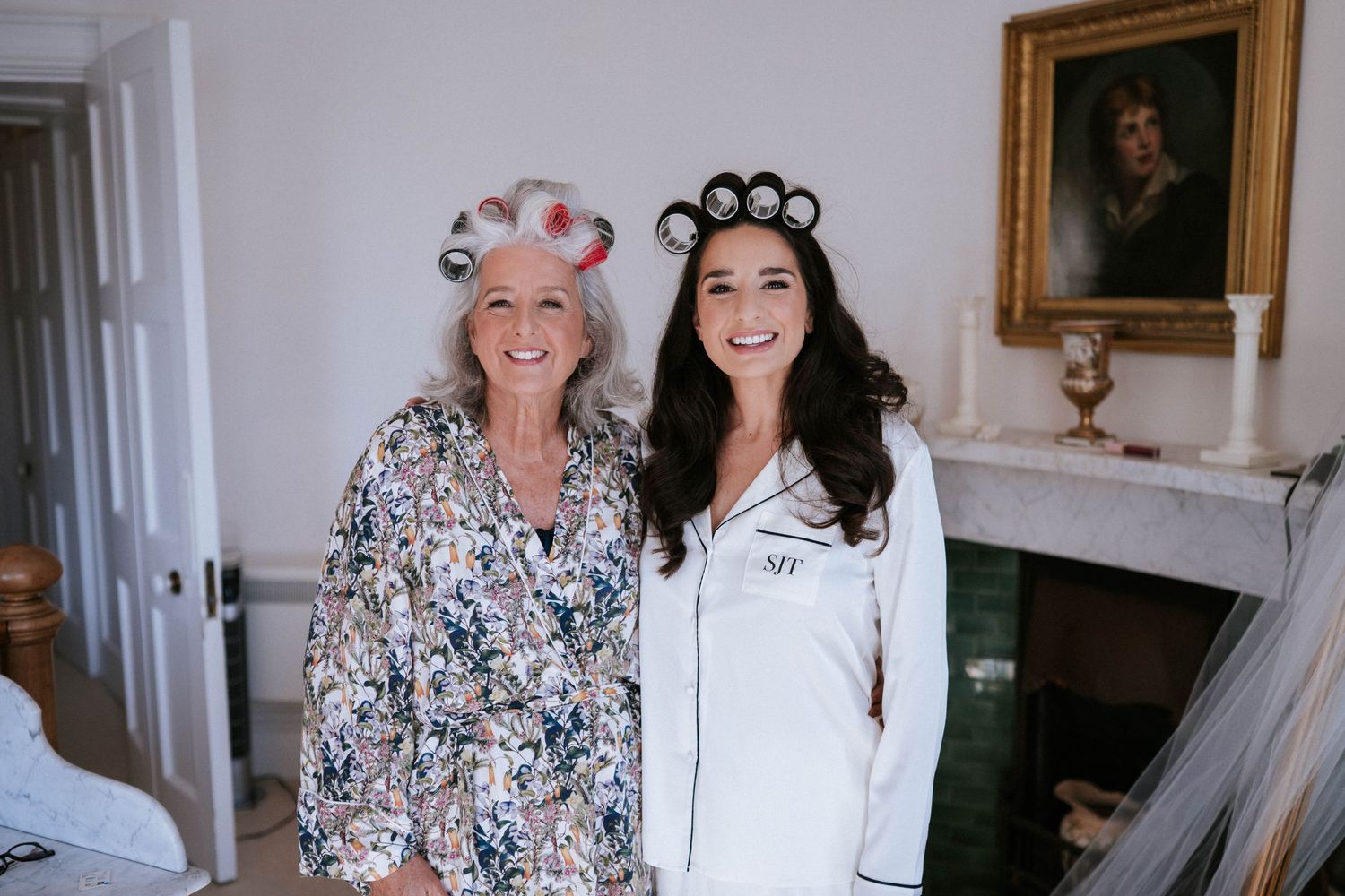 Two people wearing hair rollers and robes share a joyful moment together in an elegant room with a fireplace.