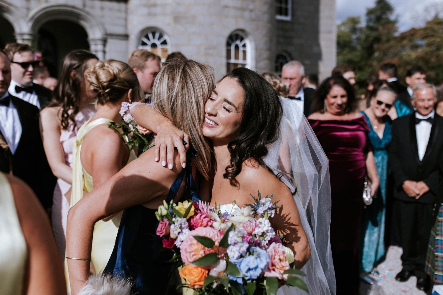 Joyful embrace between wedding guests with colorful bouquet outdoors.