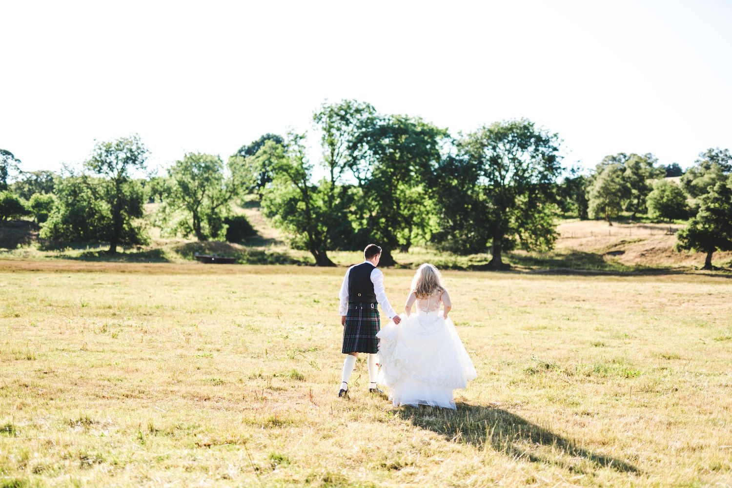 Couple walking through a sun-drenched field with scattered oak trees in the background.