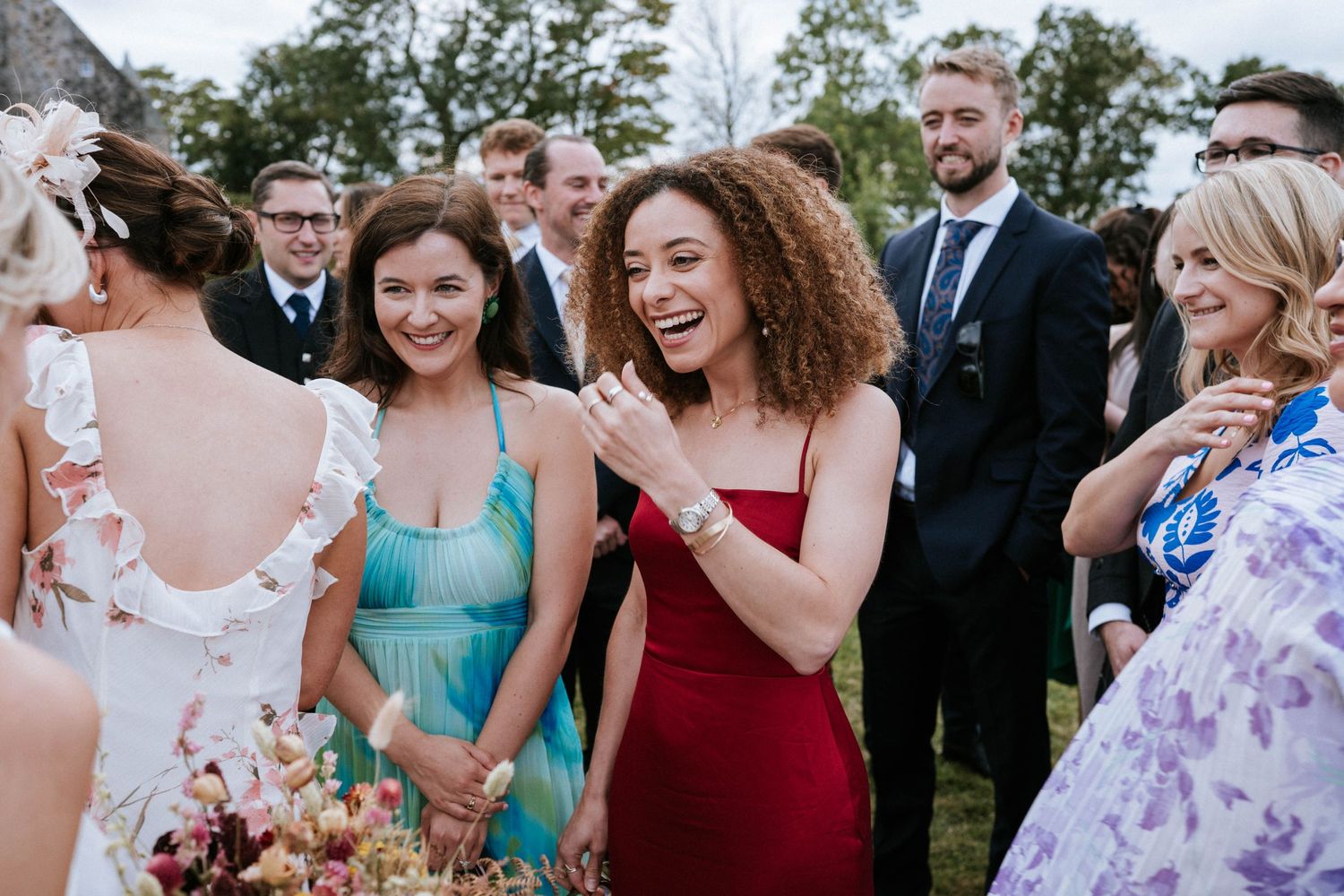 Wedding guests in colorful dresses gather outdoors for group photos on a sunny day.