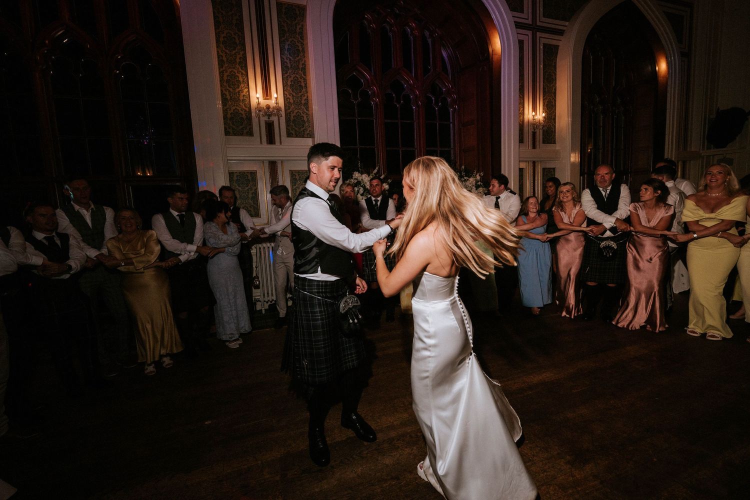A couple dances together in an elegant venue with gothic arched windows and warm lighting.