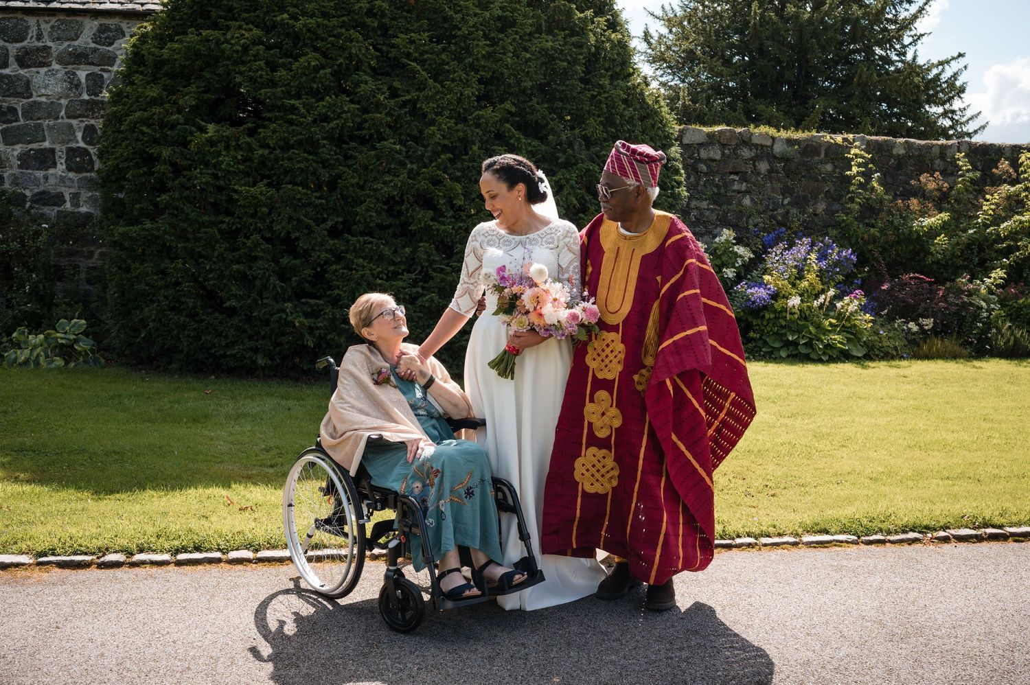 Wedding party gathered on lush green lawn outside with stone buildings in background.