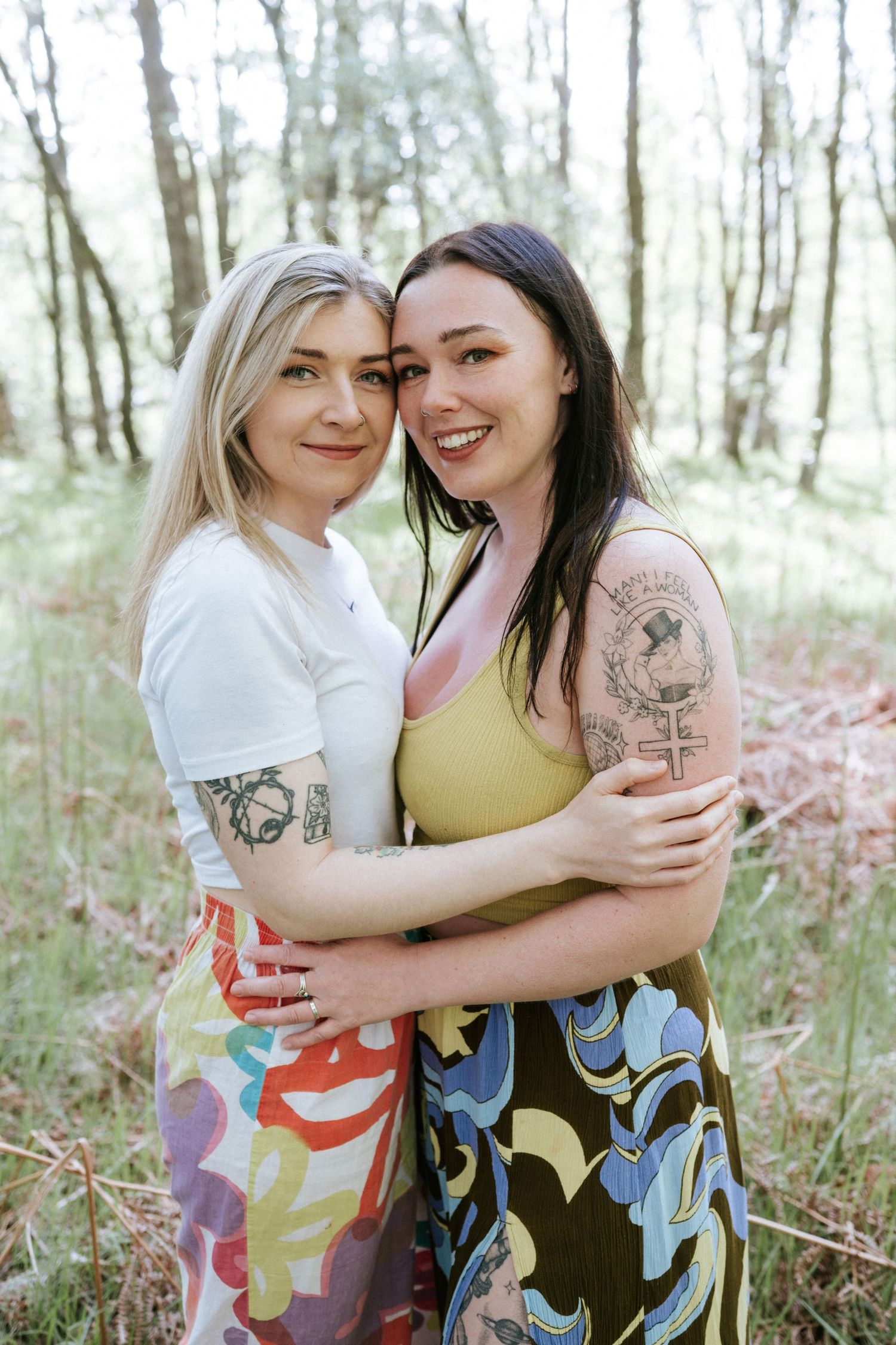 Engagement photography session Aberdeen. Two friends in colorful patterned skirts embrace and laugh together in a sunlit forest setting.