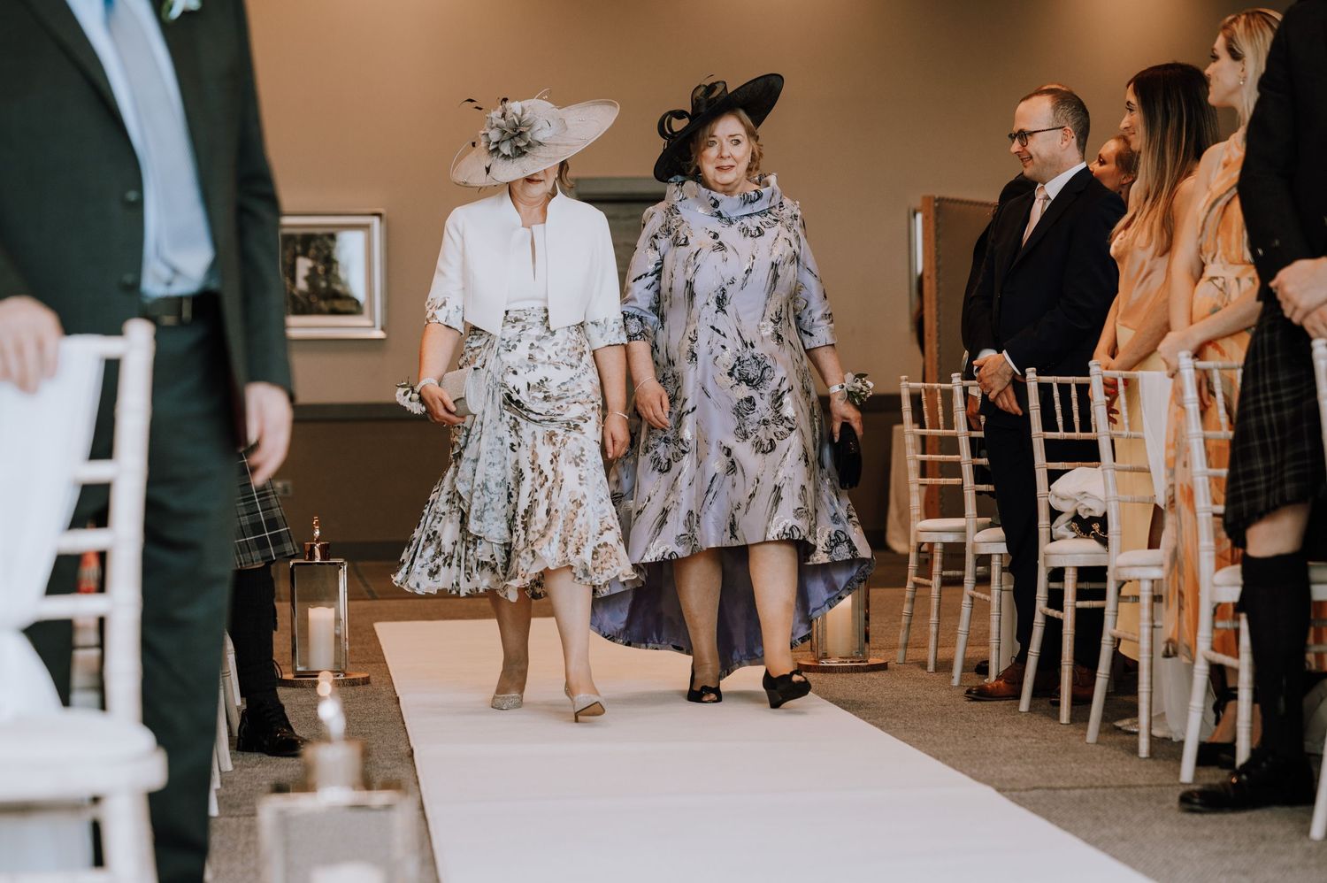 Wedding guests in formal attire walking down the aisle at an indoor ceremony.