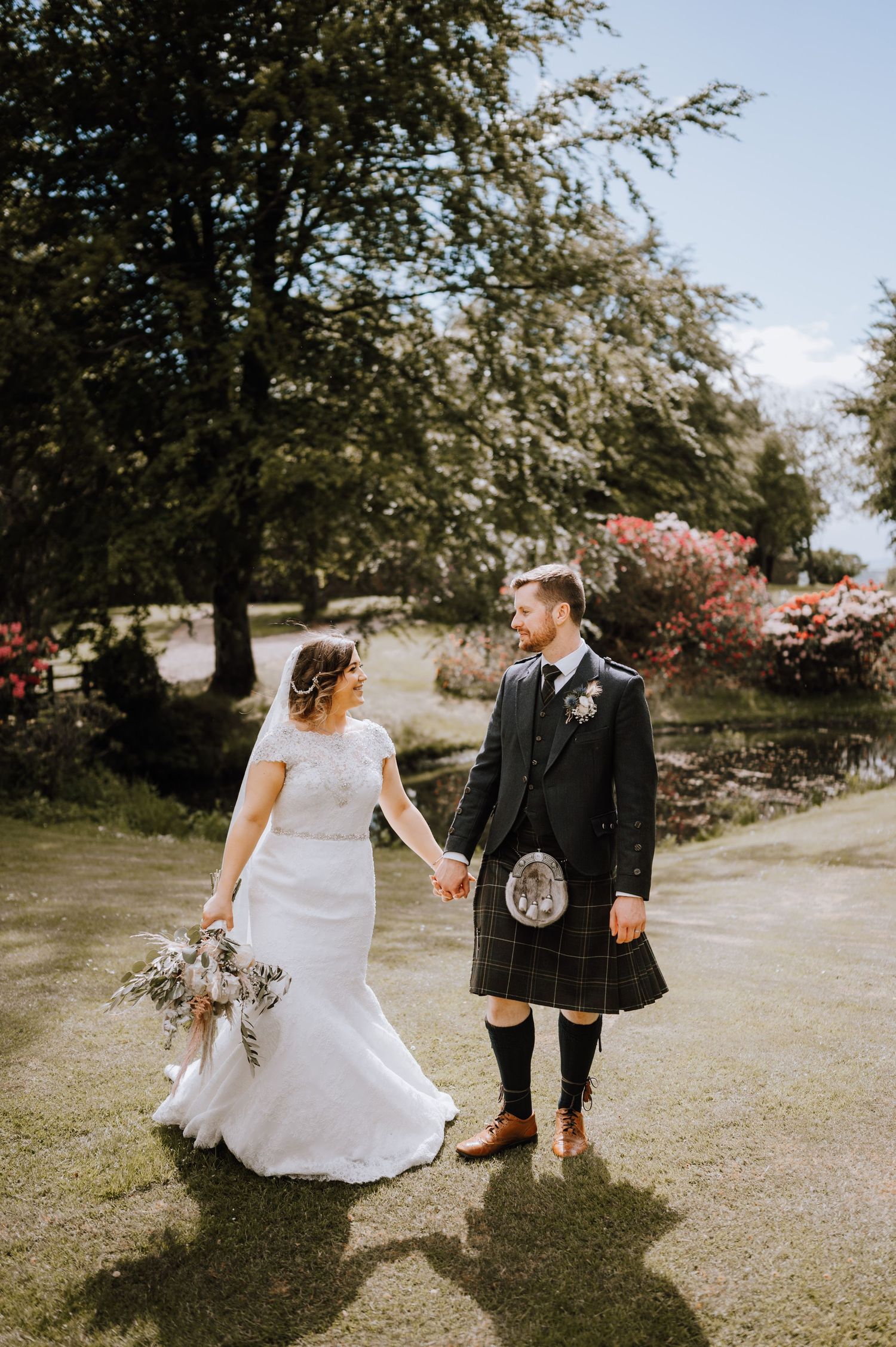 A candid wedding moment in a garden setting with the groom in traditional Scottish kilt and bride in white dress.