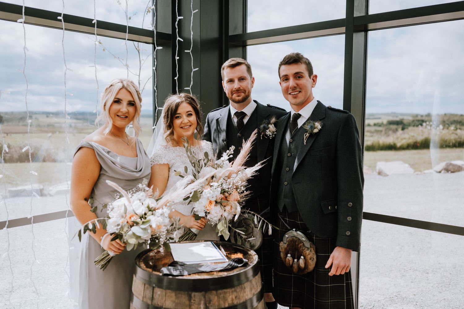 Wedding party poses with bouquets against large windows with scenic countryside views.