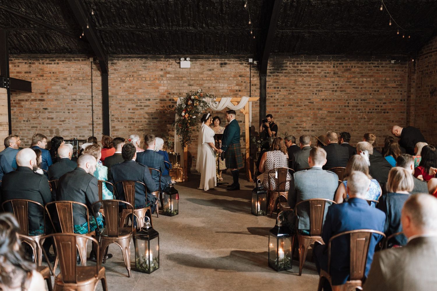 Wedding ceremony in industrial brick venue with guests seated in rows and lanterns lining the aisle.