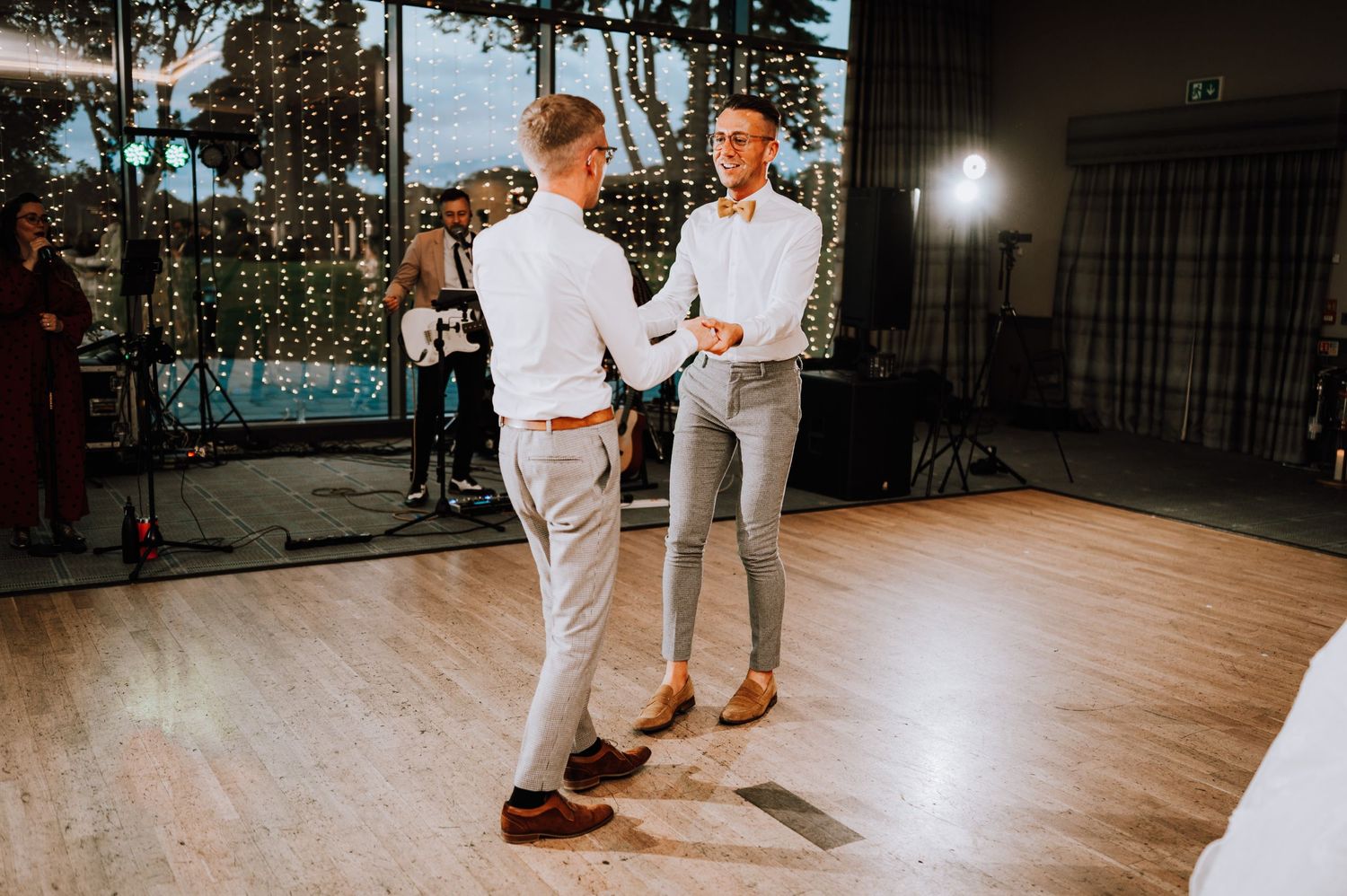 Two people in white outfits dance together on a wooden floor with string lights in the background.