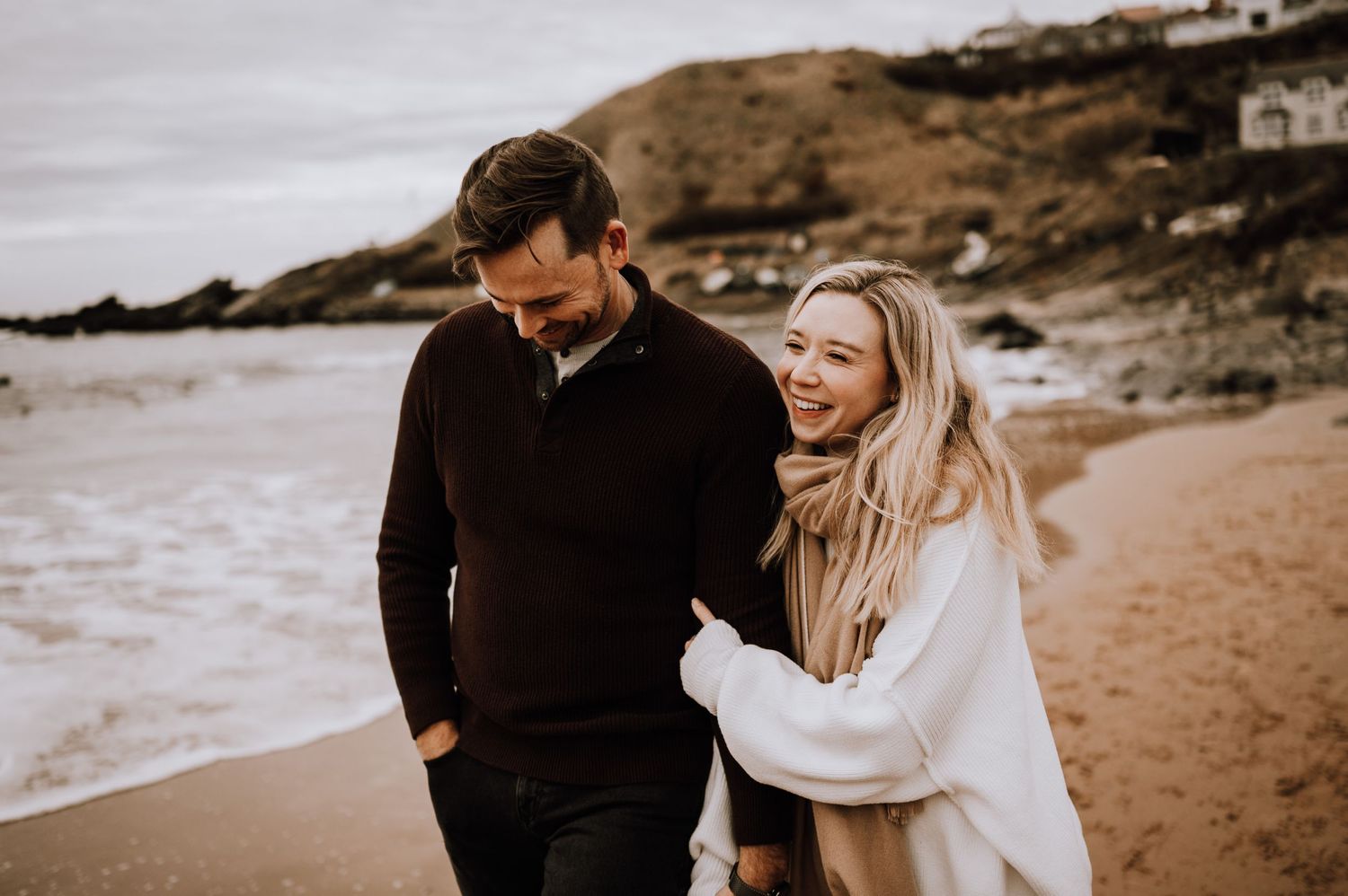 A joyful couple laughing together during their beach photoshoot with coastal scenery.
