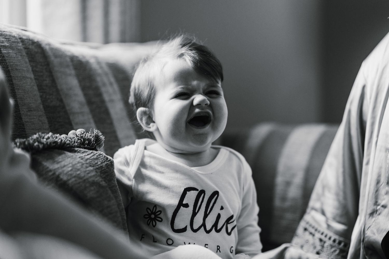 A black and white portrait of a laughing baby wearing an Ellie shirt on a couch.