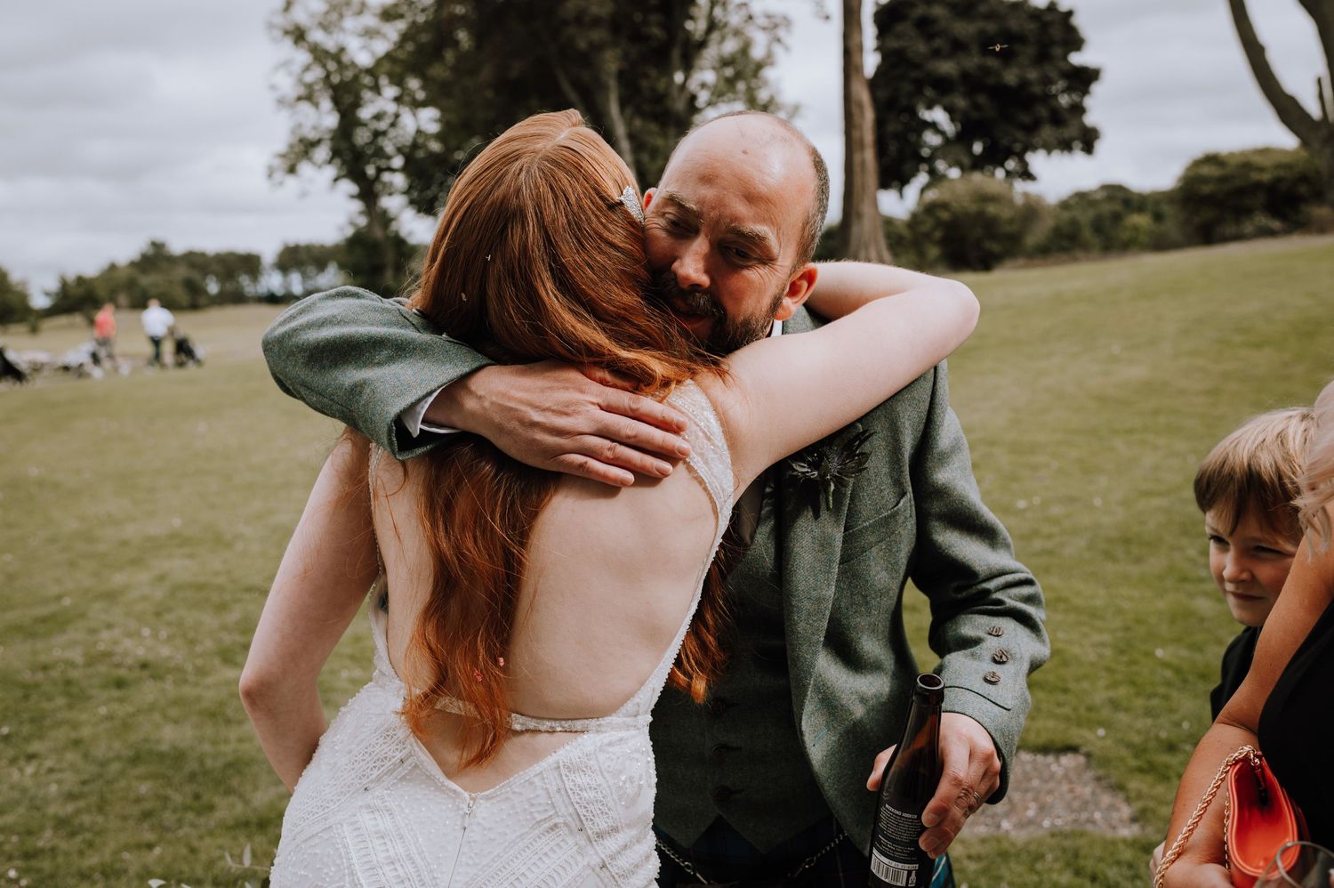 An emotional embrace during an outdoor wedding celebration, with long red hair flowing in the sunlight on a grassy field.