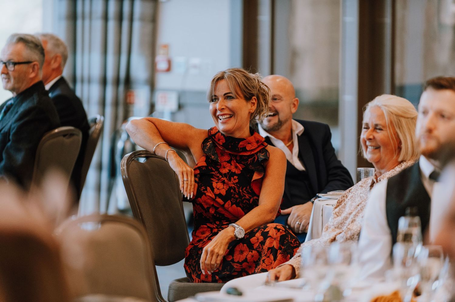 Wedding guests in formal attire share laughter during reception speeches.