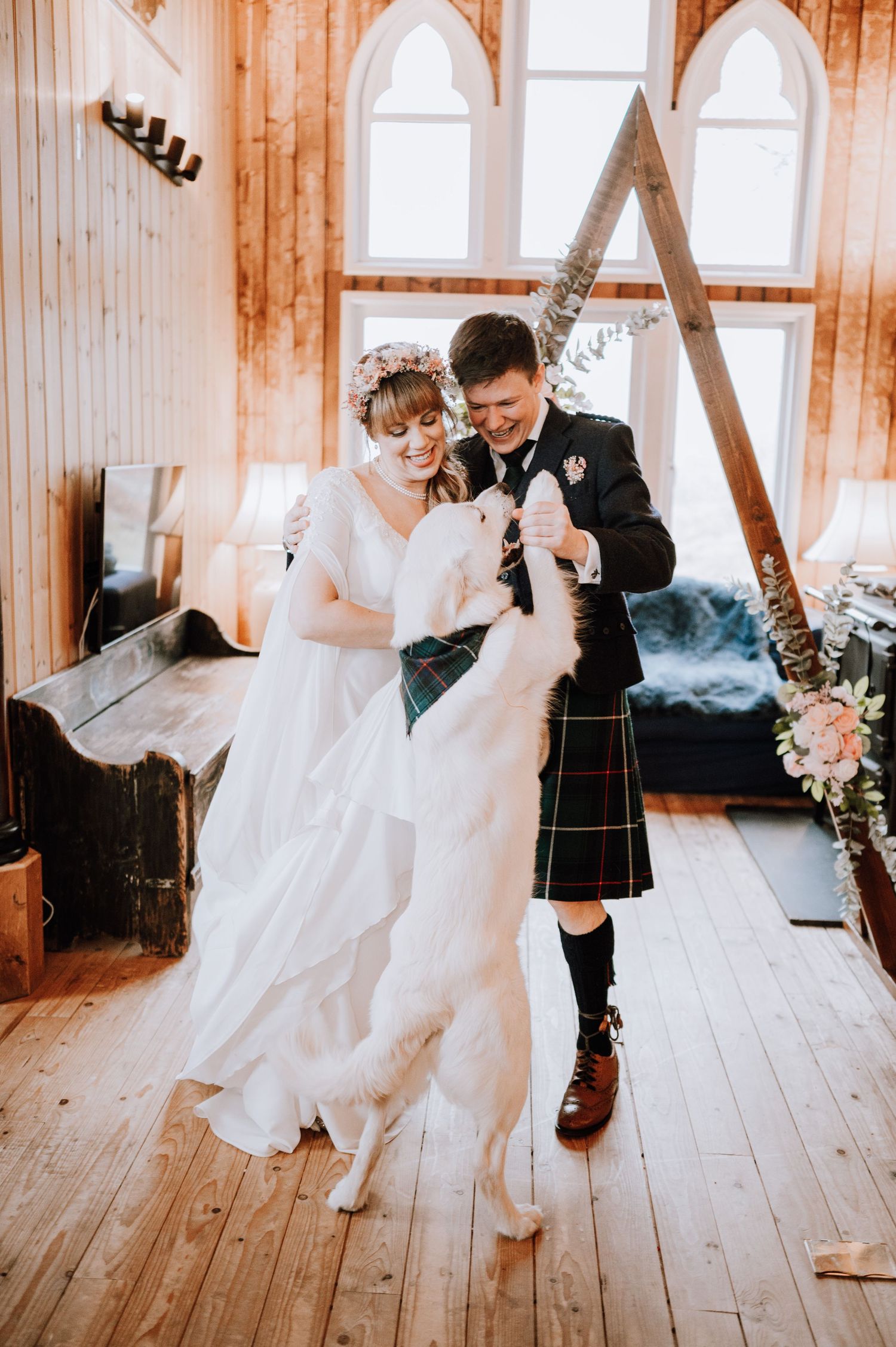 Couple dances with their dog under triangle arch in rustic wooden venue with arched windows.