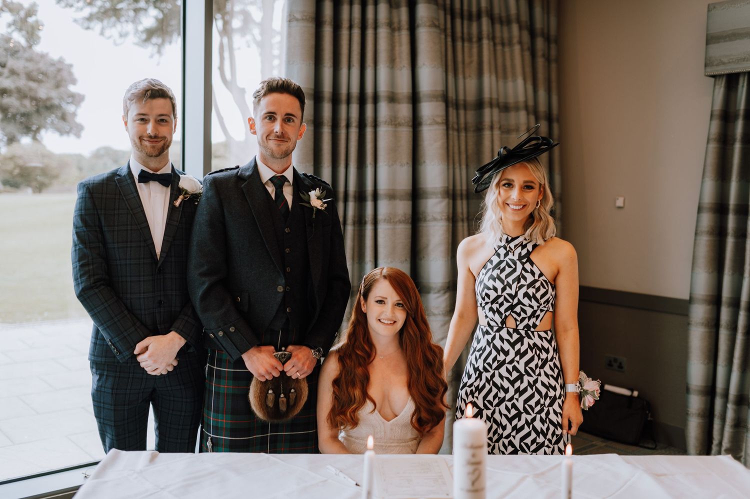 Wedding party posing together in formal attire near large windows.