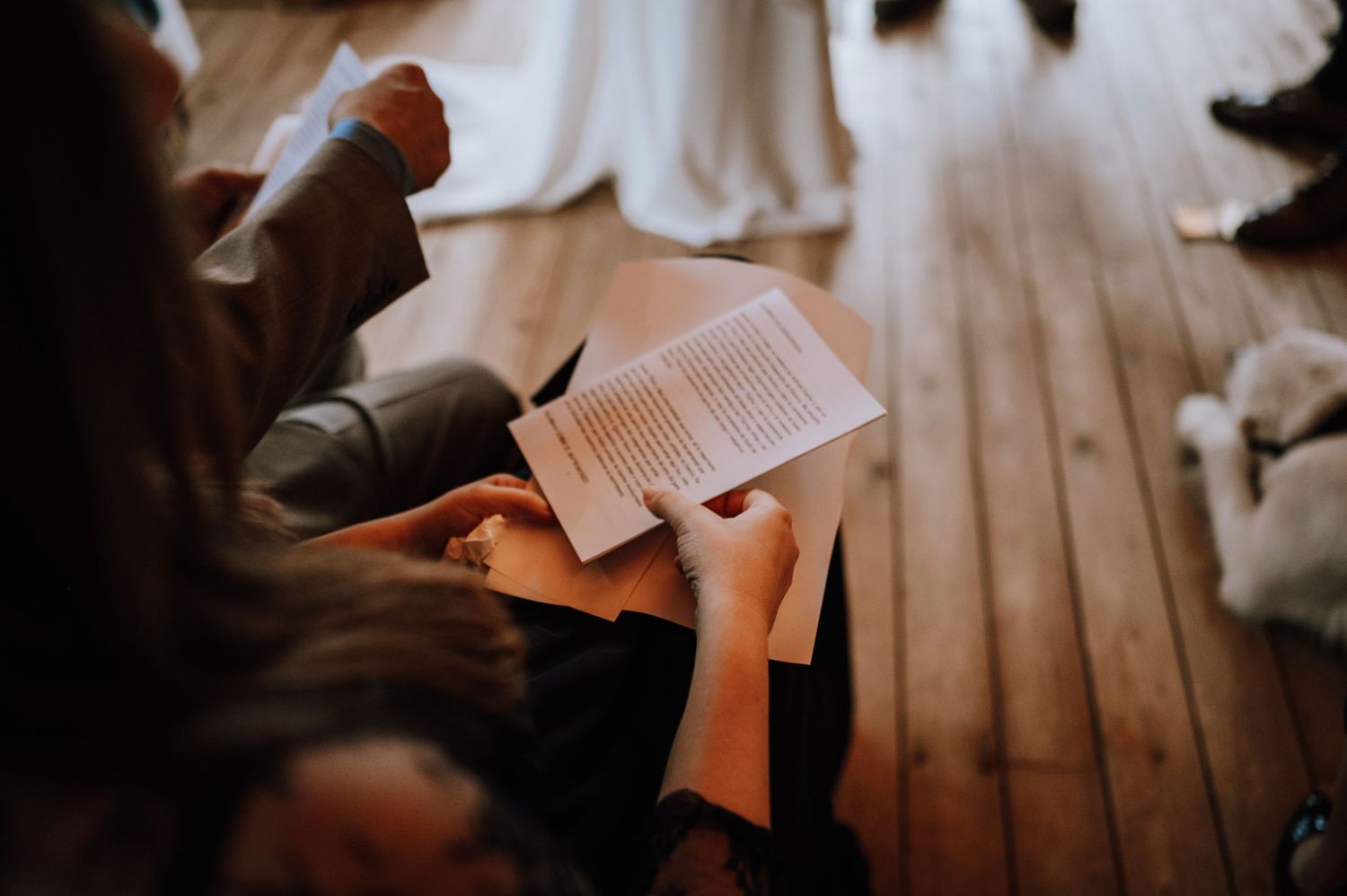 A person reads from an open book while sitting on wooden floorboards in warm lighting.