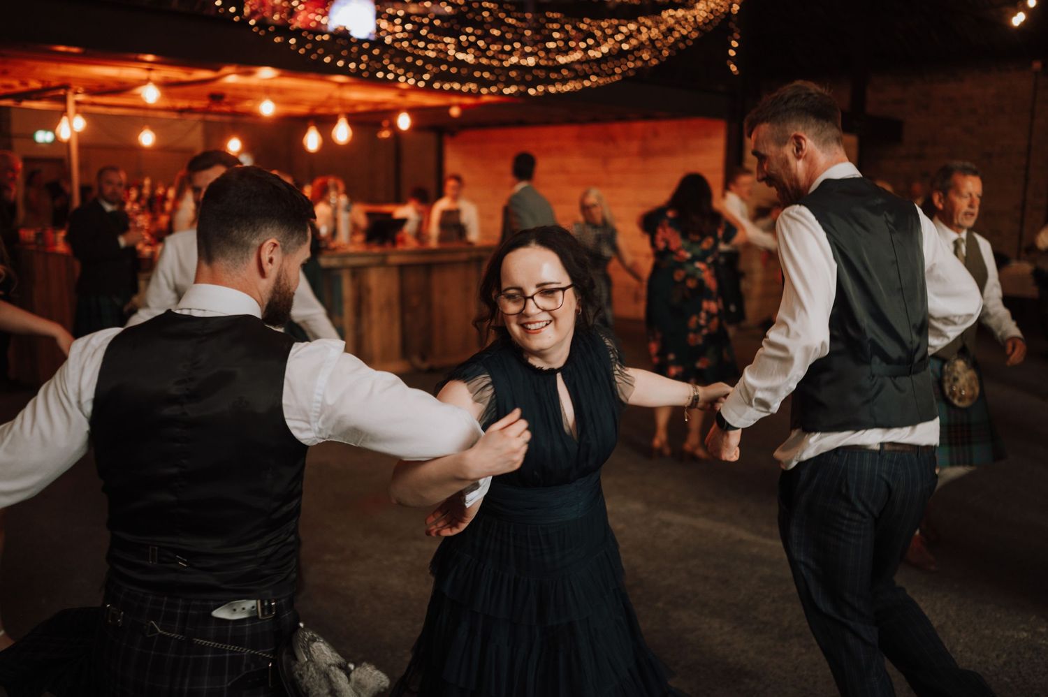 Dancing guests enjoy a festive celebration in a rustic venue with brick walls and string lights.